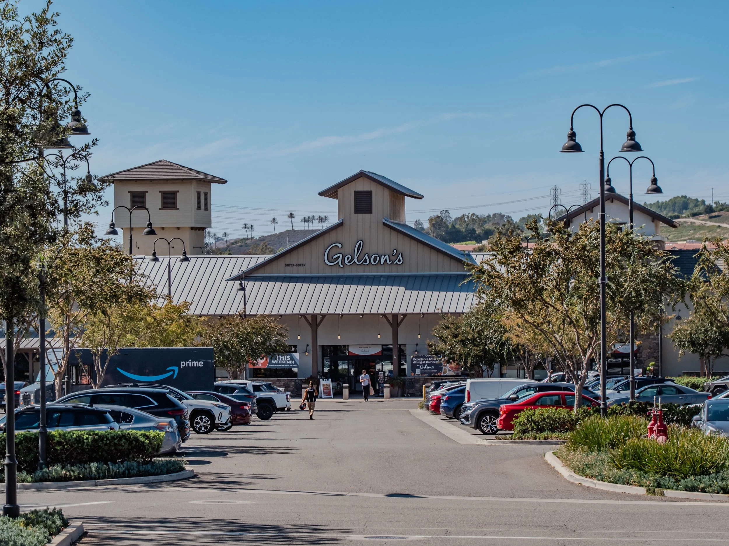 Parking lot in front of Gelson's supermarket with cars parked and a few people walking toward the entrance, surrounded by trees and street lamps under a clear blue sky.