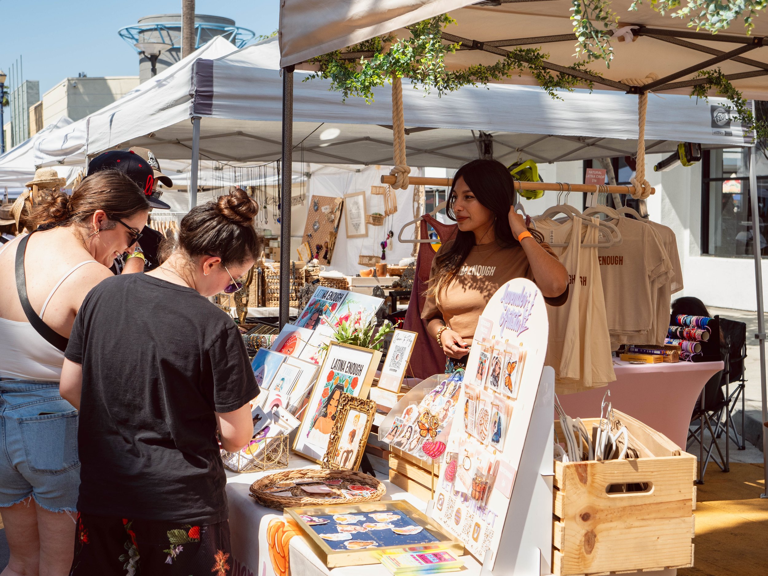 A woman with long dark hair wearing a tan shirt browsing jewelry at an outdoor market booth, as two other women look at items on the table.