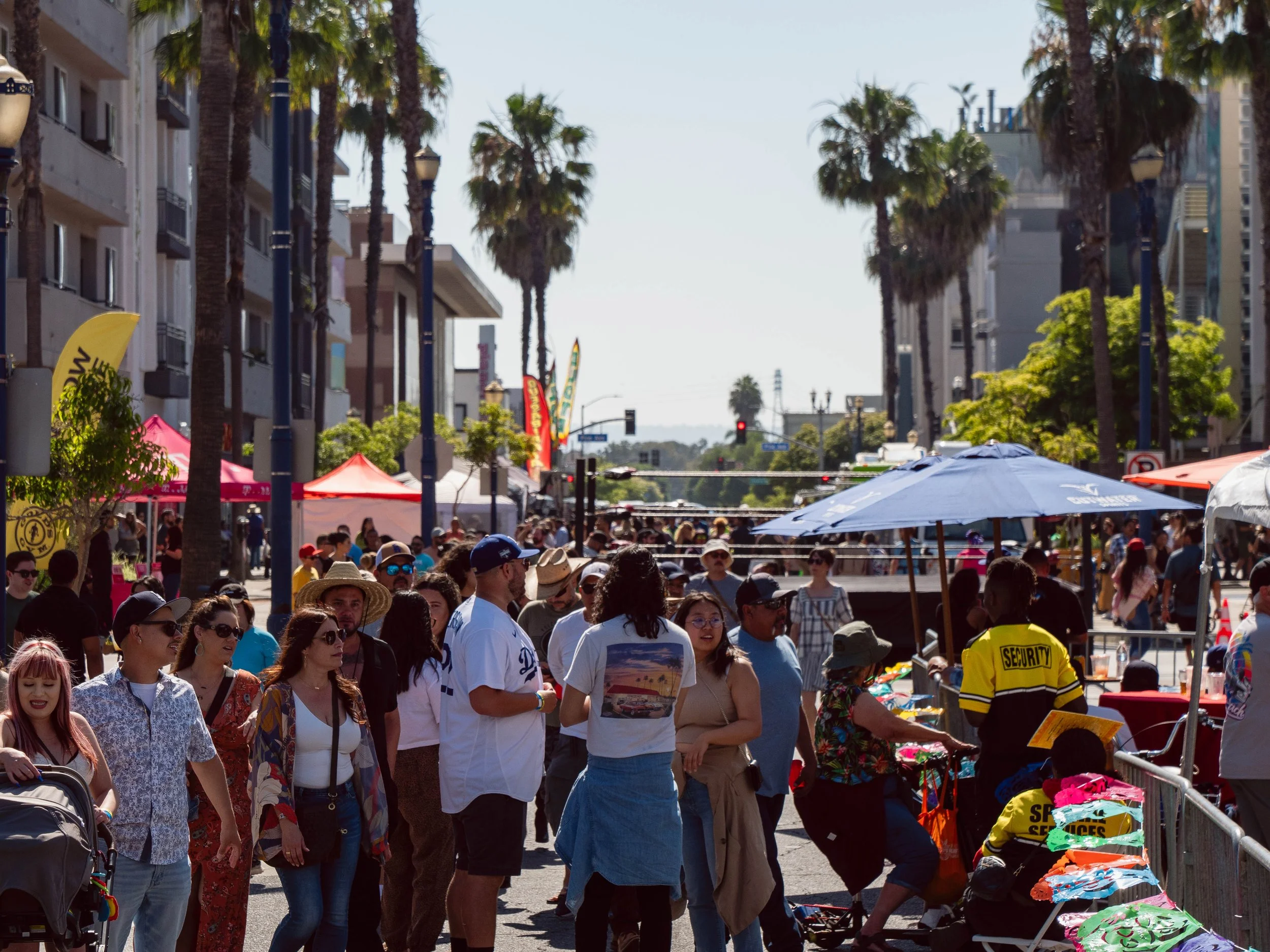 Crowd of people at an outdoor market or festival with tents and umbrellas, palm trees, and buildings in the background.