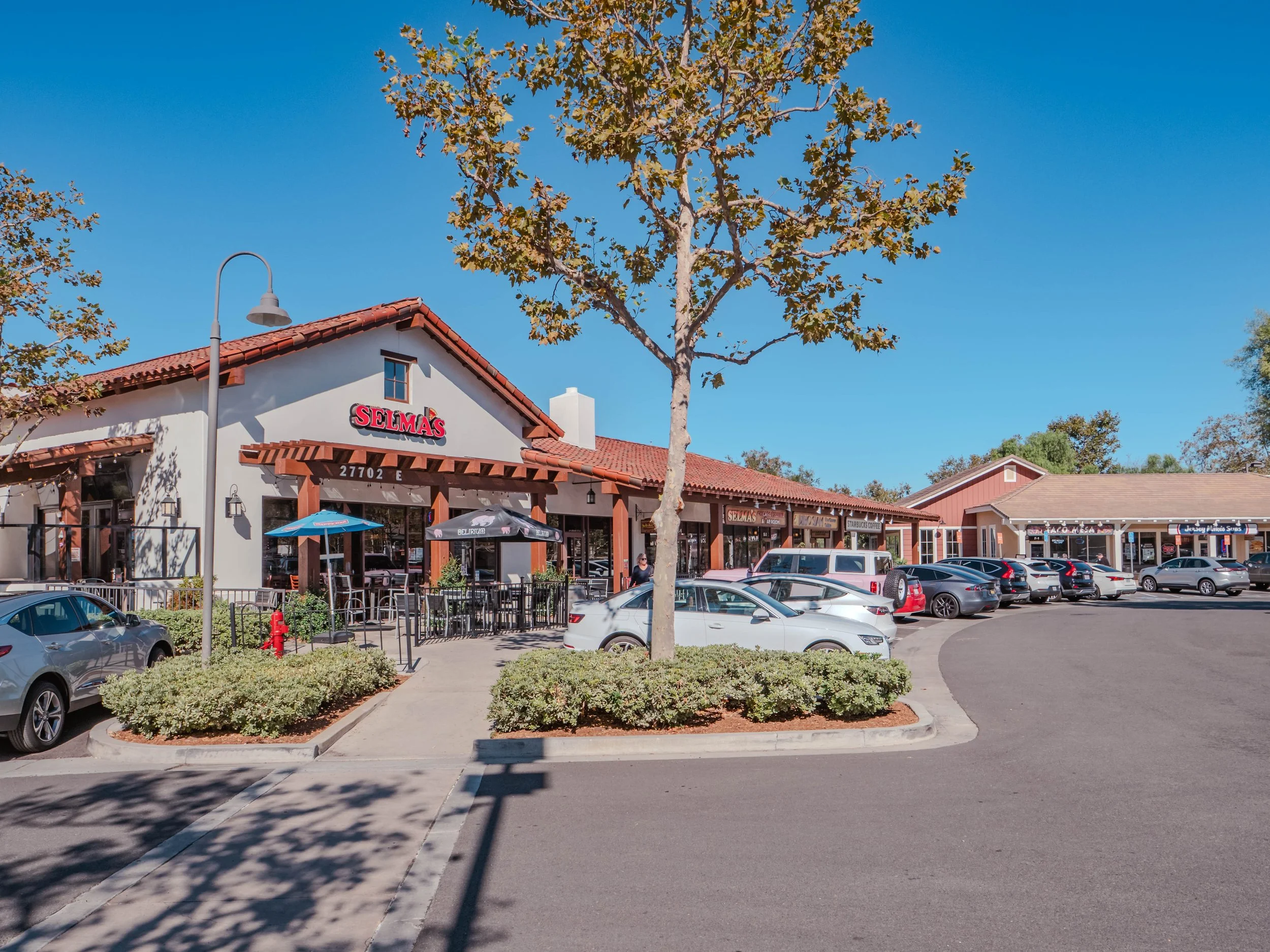 A shopping plaza with a restaurant called Selma's, outdoor seating with umbrellas, and several parked cars. The scene includes a sidewalk, a tree in the center, and other shops in the background under a clear blue sky.