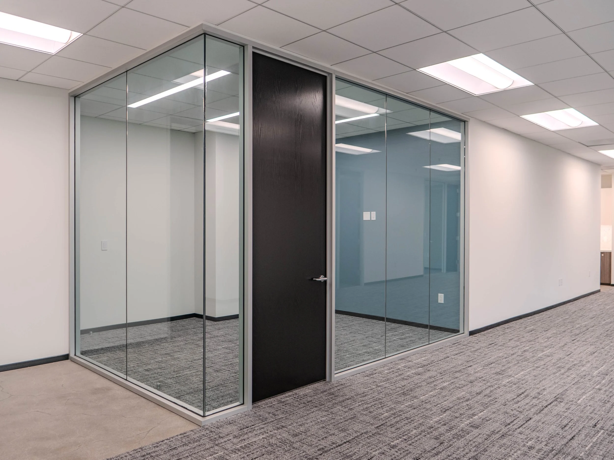 Modern office space with glass enclosed meeting room, featuring a black door and gray carpeted flooring.