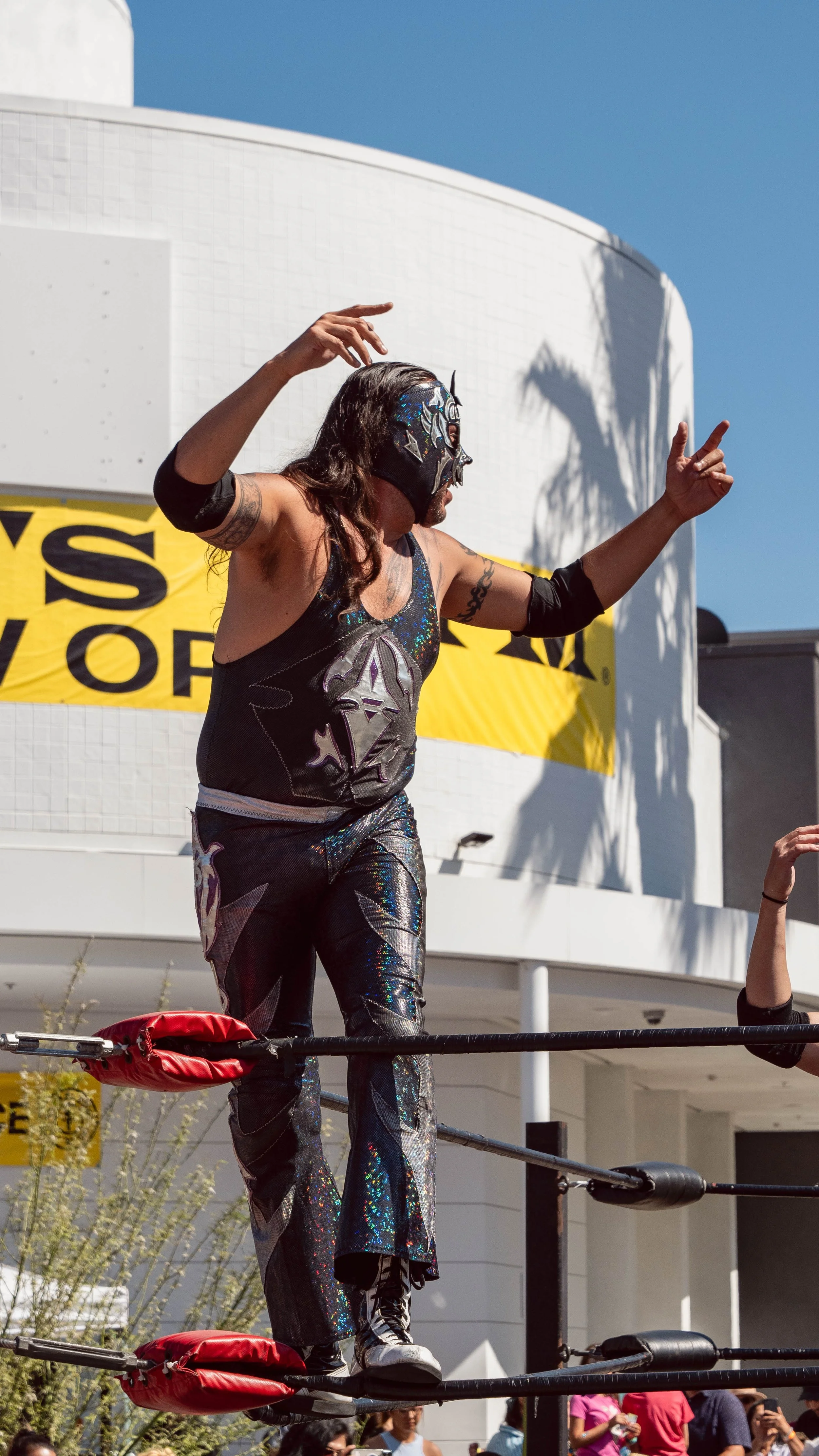 A masked wrestler standing on the top rope of a wrestling ring, gesturing with both hands during an outdoor event in front of a white building and yellow banner.