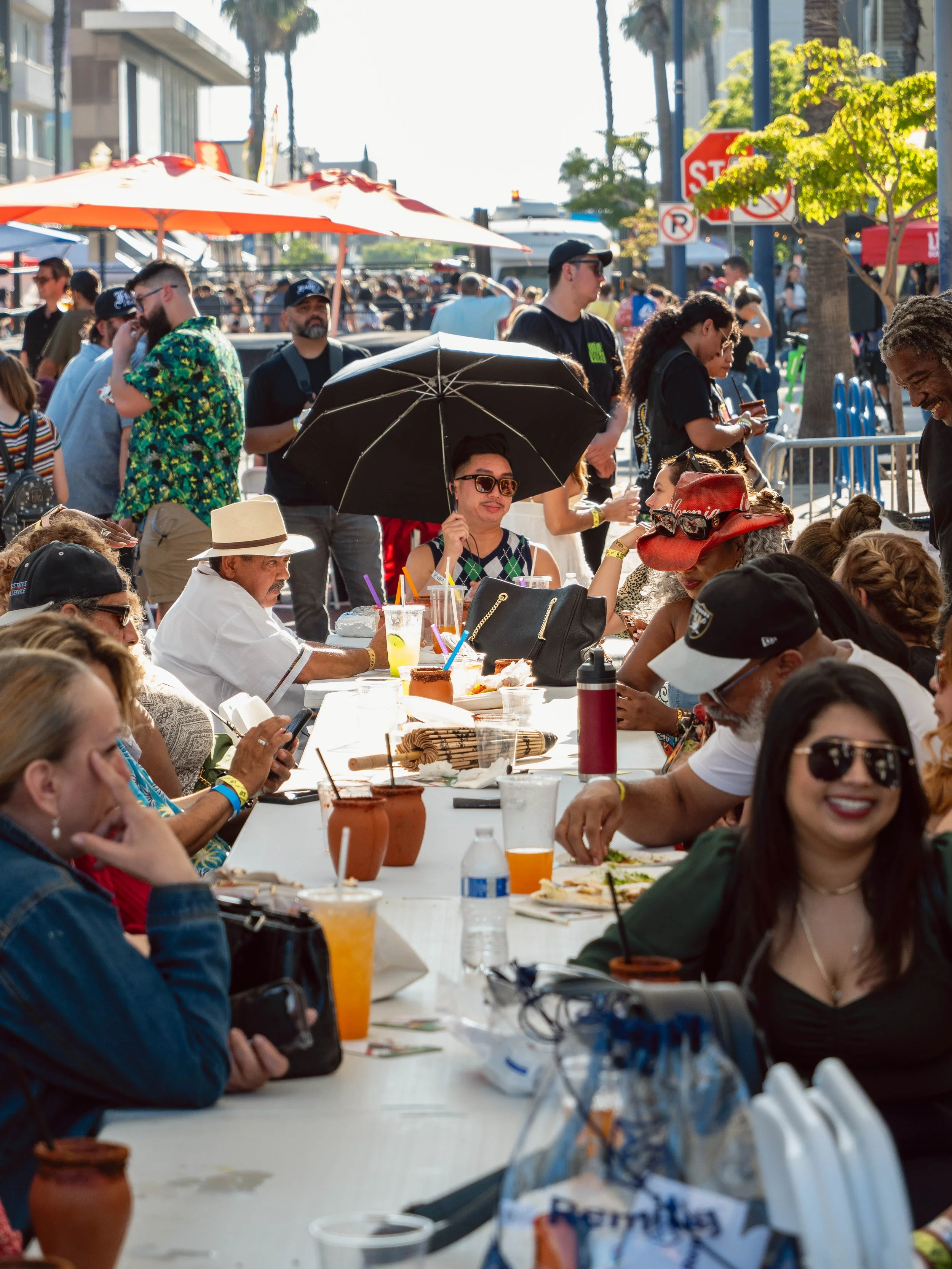 People enjoying a sunny outdoor event at a long white table with food and drinks, some wearing hats and sunglasses, in a vibrant street scene with tents, umbrellas, and trees.