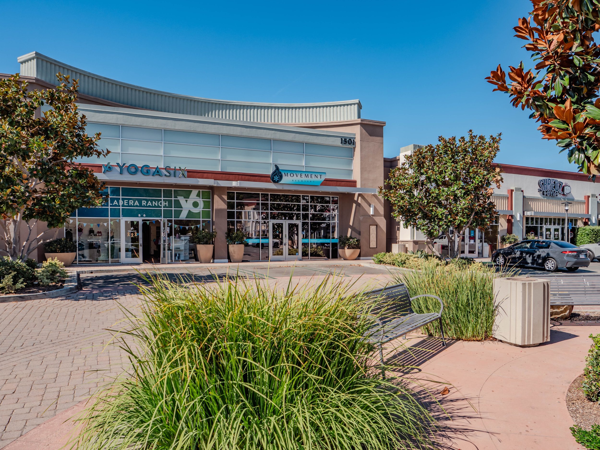 Shopping plaza with storefronts, benches, trees, and parked cars under a clear blue sky.