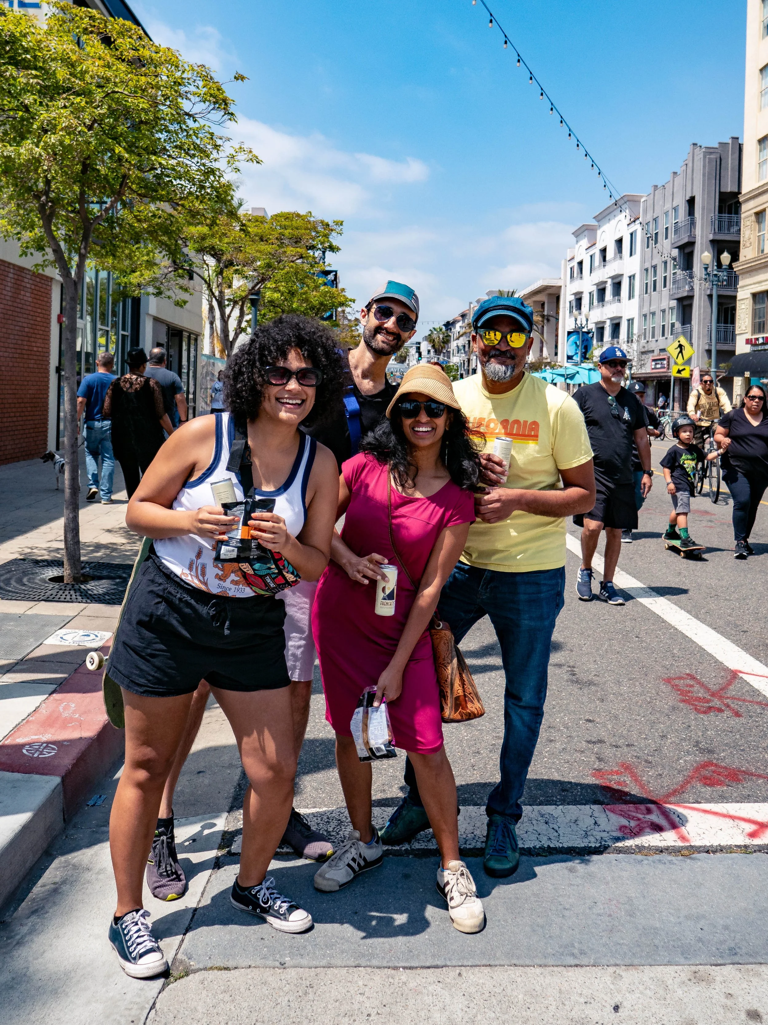 Four people smiling and posing for a photo on a sunny street, with shops, buildings, and other pedestrians in the background.