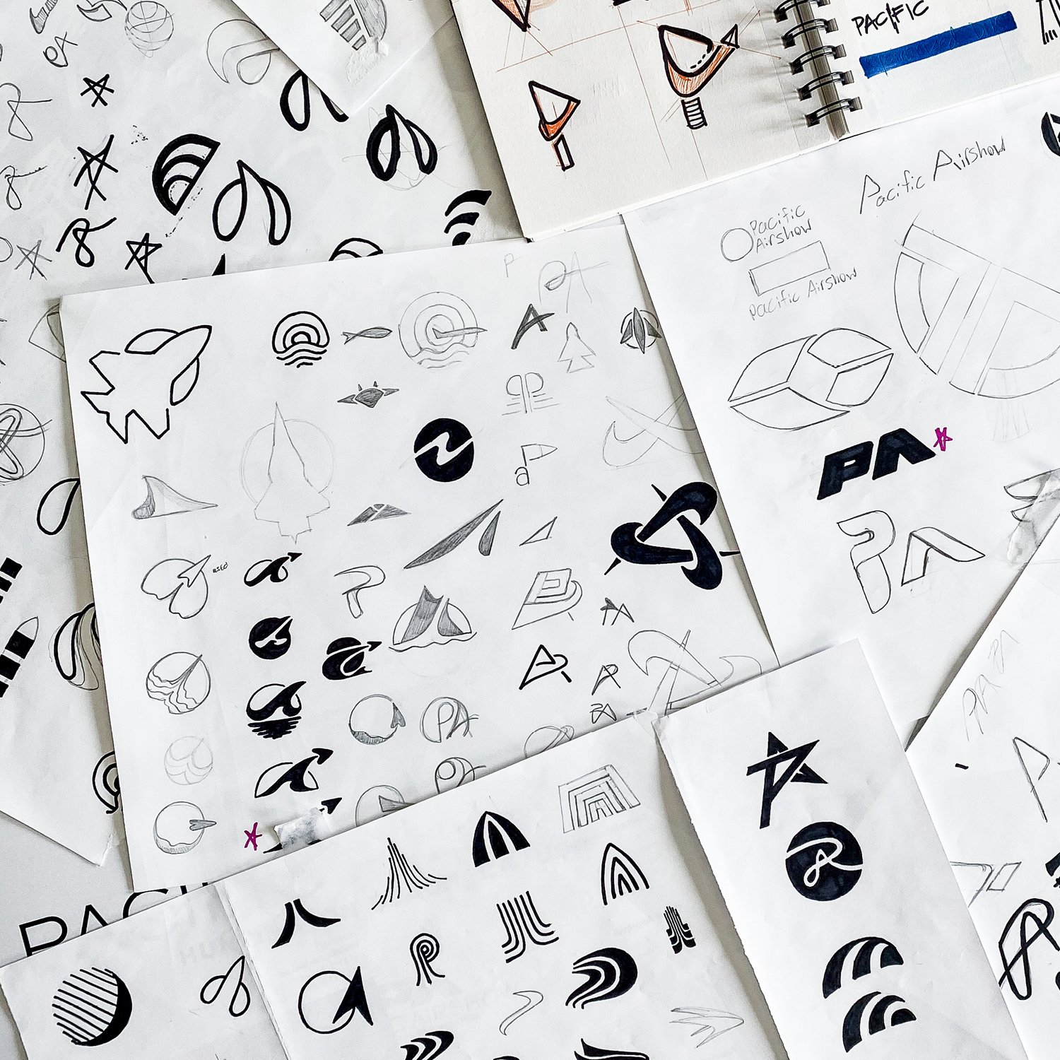 Several sheets of paper with various logo sketches and designs, some featuring geometric shapes, stylized symbols, and abstract forms.