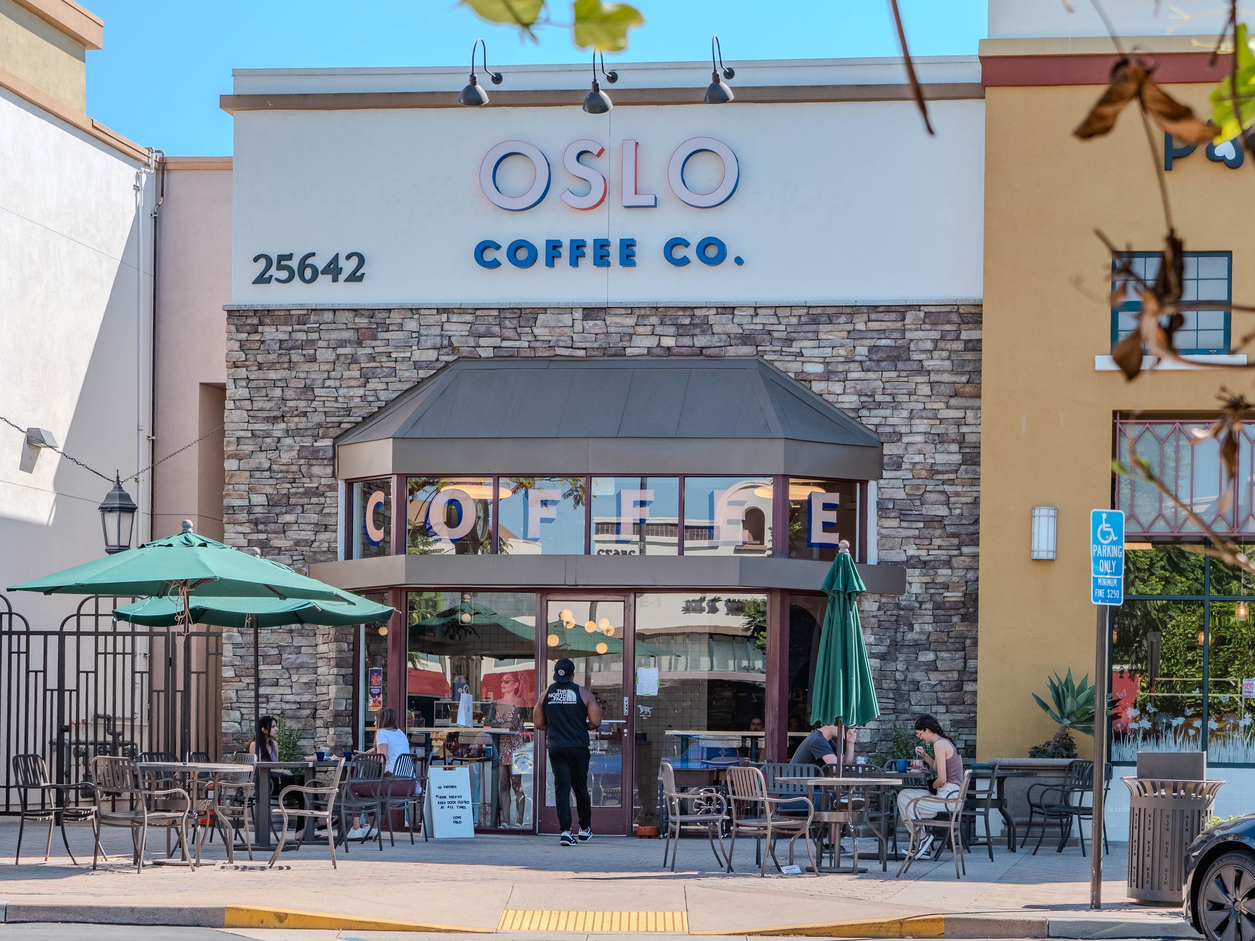 Exterior view of Oslo Coffee Co., a café with outdoor seating, umbrella shade, and people sitting at tables, located at 25642 with a stone façade and sign.