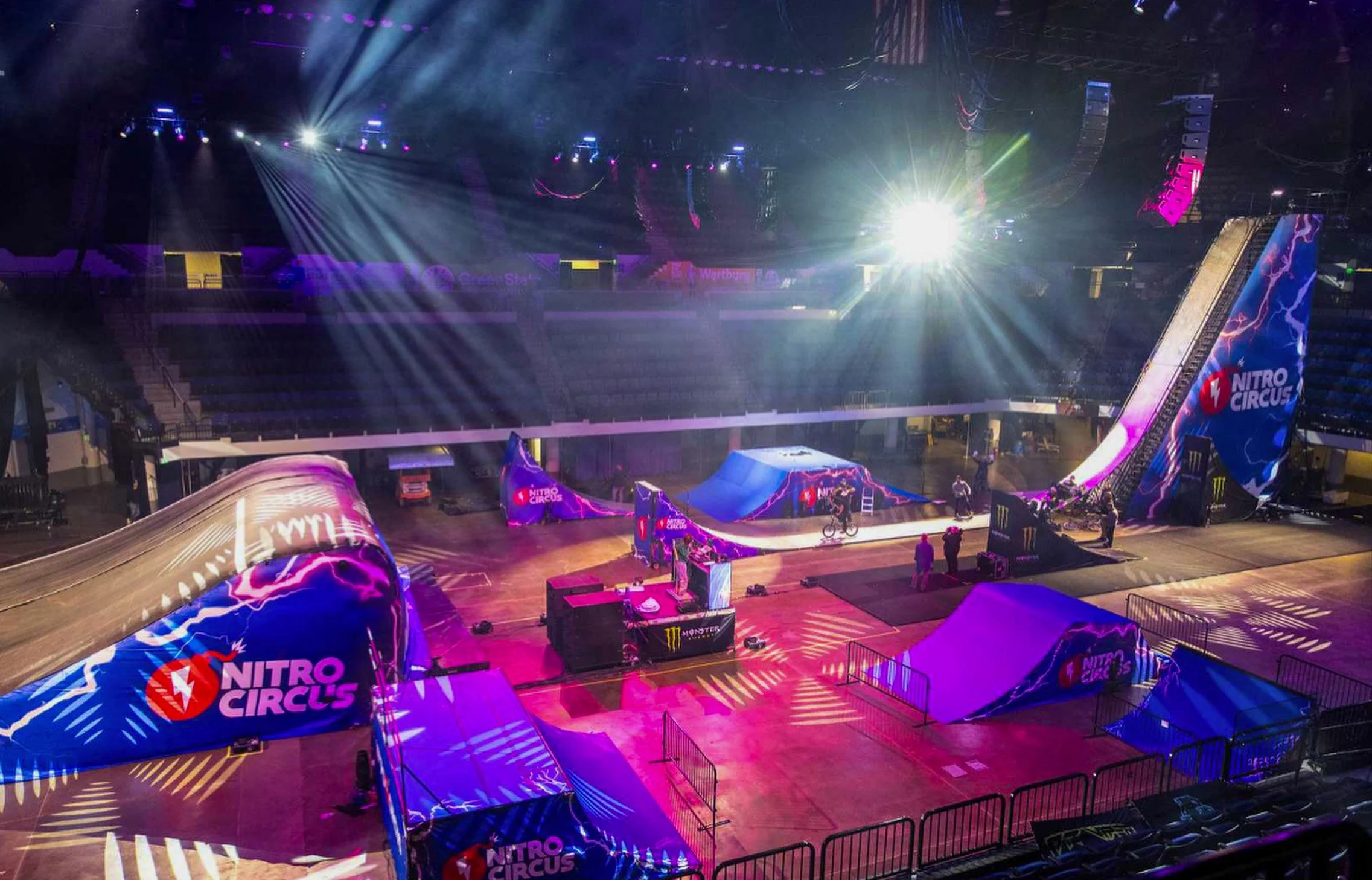 Indoor dirt bike stunt arena with ramps, airbag landing, and Nitro Circus branding, illuminated by colorful lighting.
