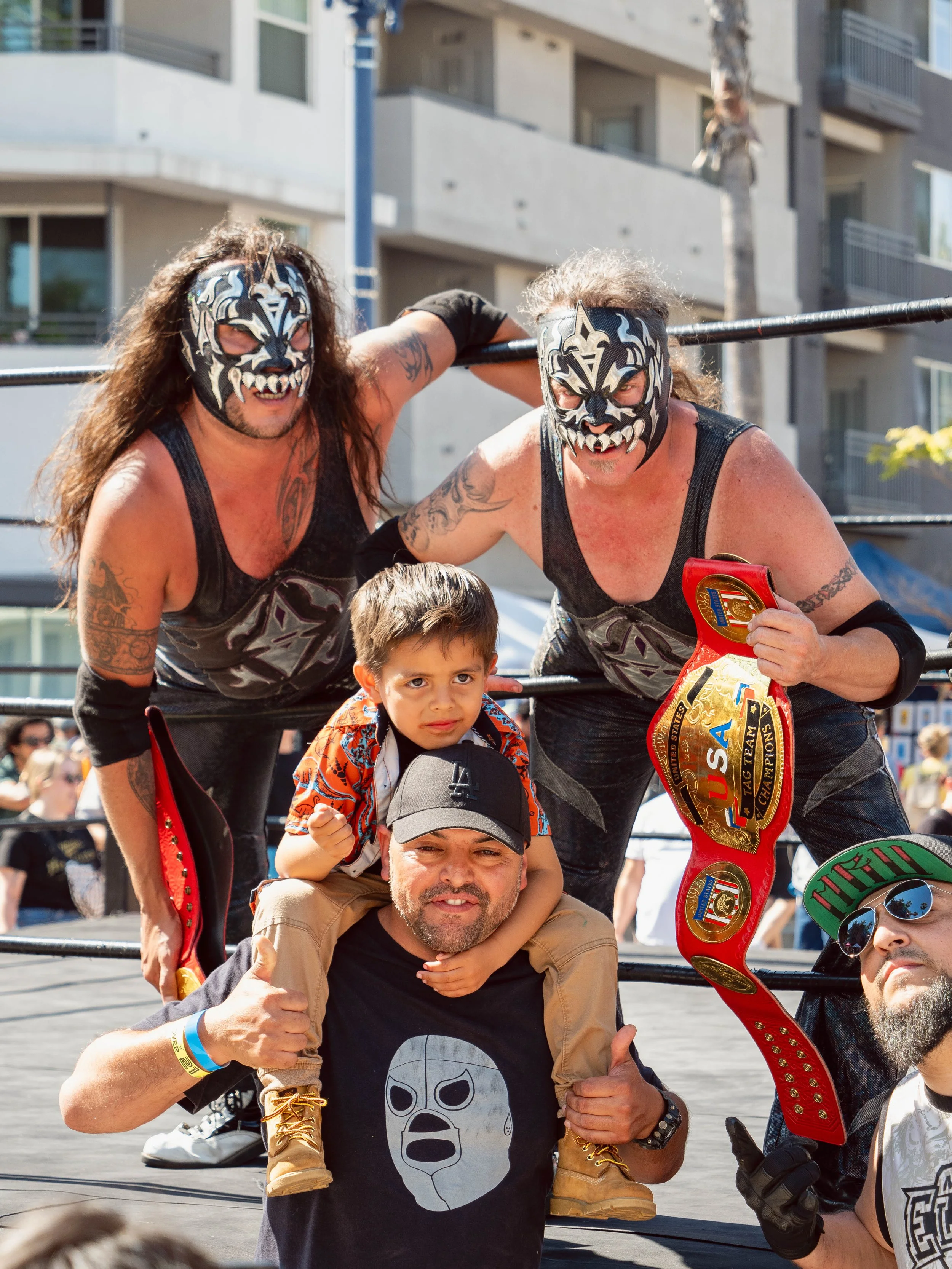Two wrestlers in black masks and costumes posing in a wrestling ring, holding a championship belt. A young boy and two men are standing in front of them, one man is giving a thumbs-up, and the boy is sitting on his shoulders.