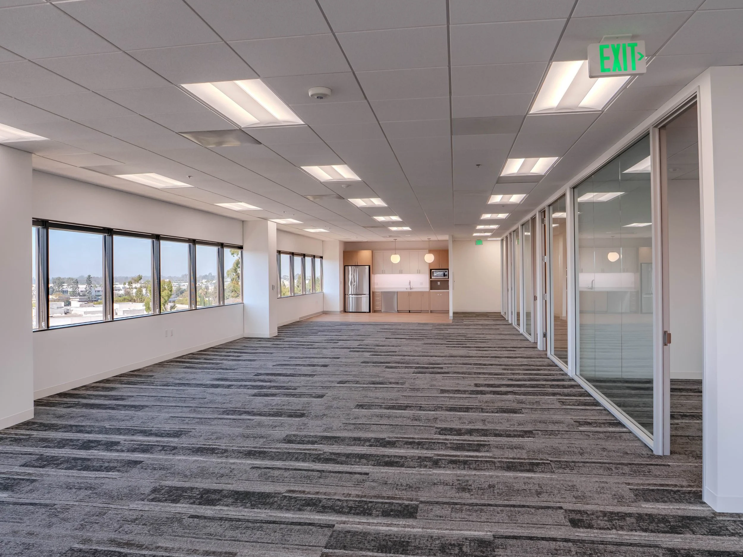 Modern office space with large windows, carpet flooring, glass partitions, a mini kitchen area with refrigerator and microwave, and an exit sign on the ceiling.