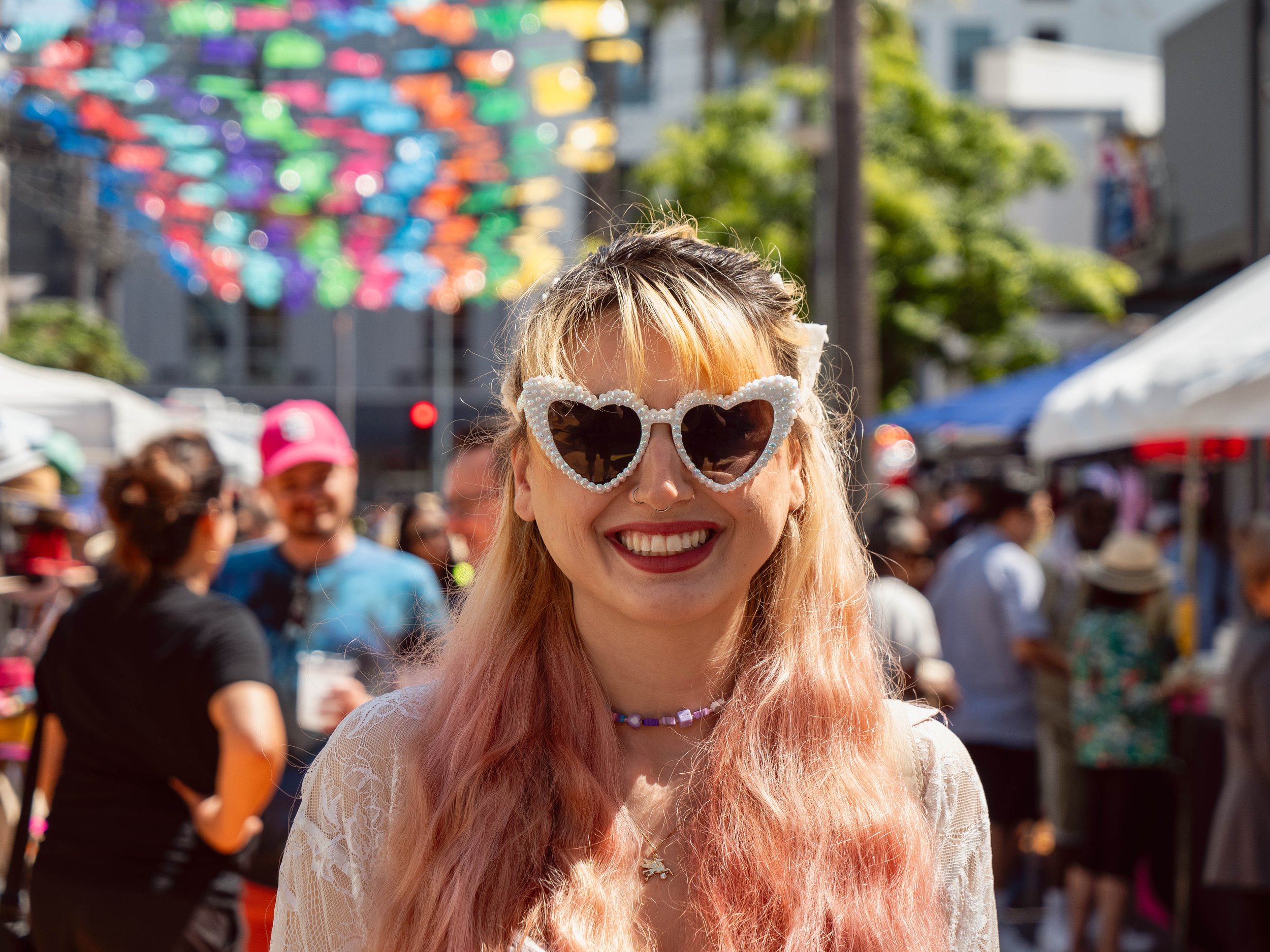 A woman smiling, wearing heart-shaped sunglasses, in a crowded outdoor festival or market on a sunny day, with colorful decorations overhead.