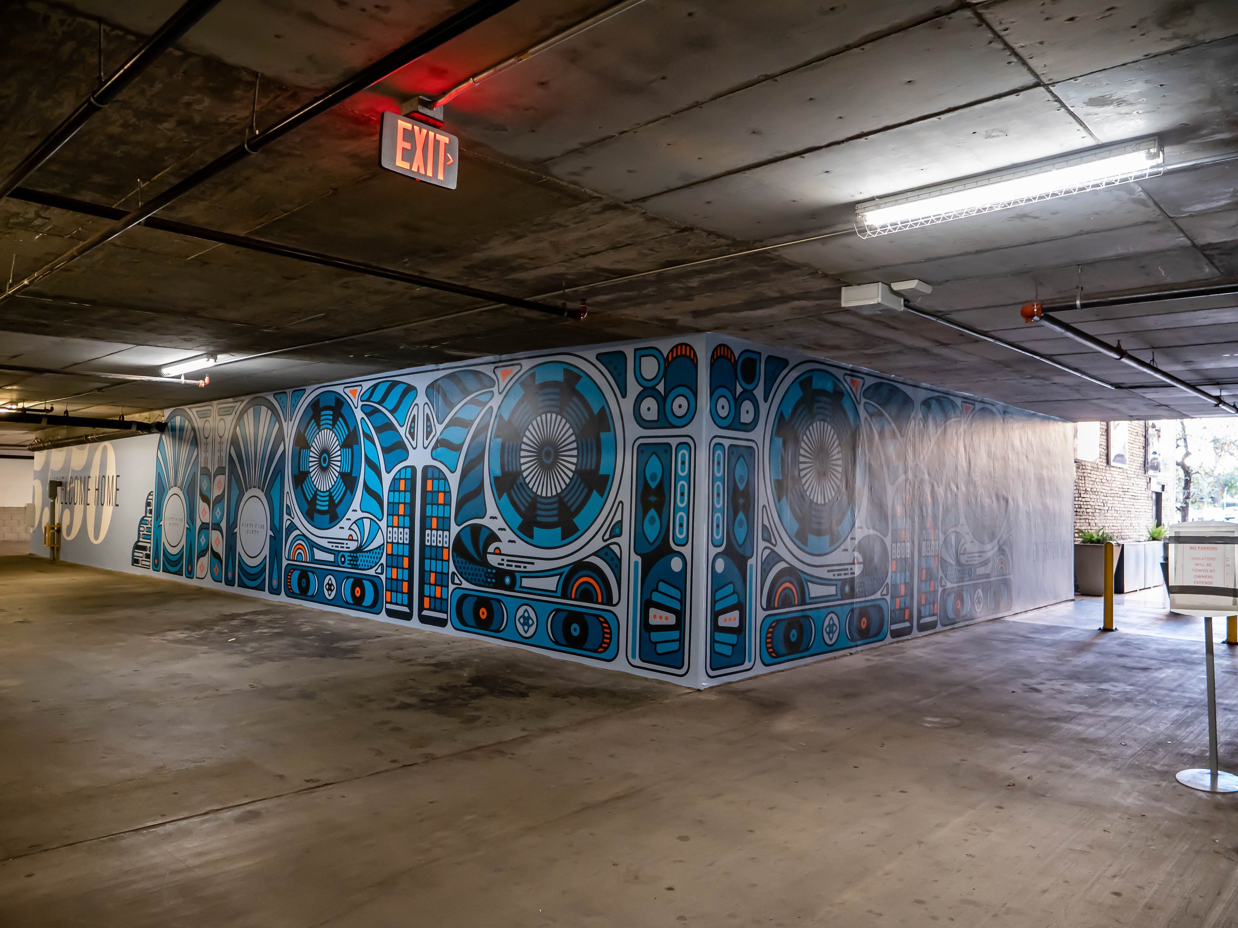 Colorful abstract mural in an underground parking garage with concrete ceiling and floor, illuminated by fluorescent lights, and a visible exit sign.