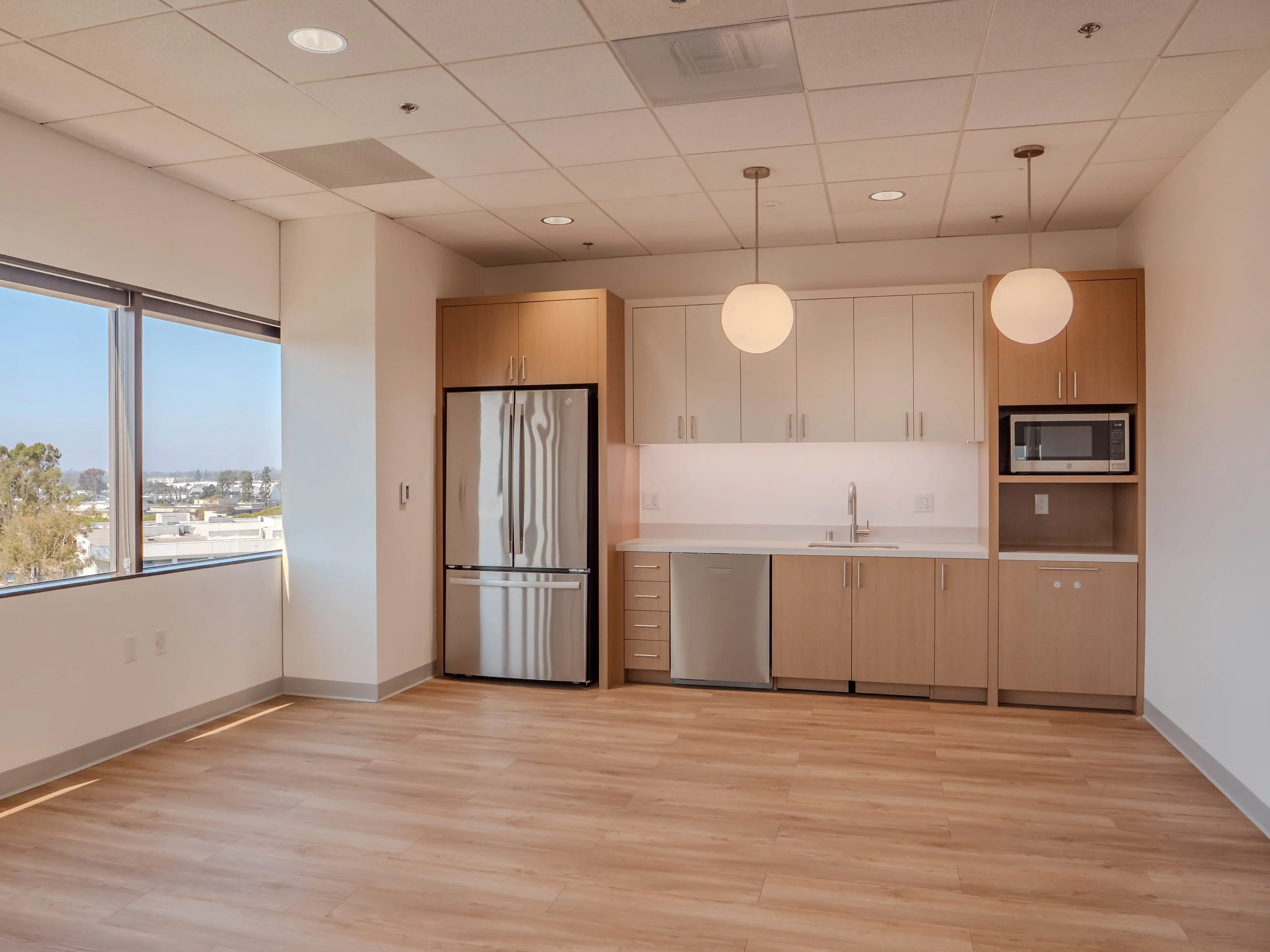 An empty kitchen with light wood cabinets, stainless steel appliances, a white countertop, and two hanging pendant lights. Large windows on the left offer a city view.