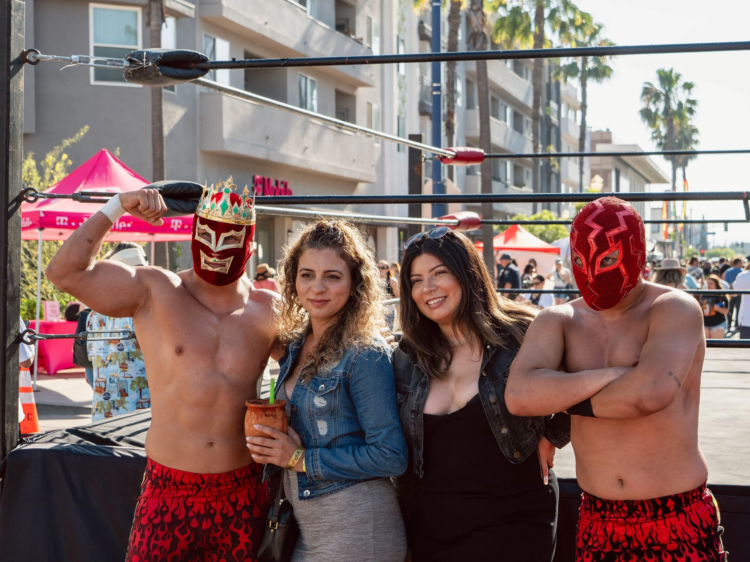 Two men wearing red lucha libre masks and red flame-patterned shorts pose with two women in casual clothing at an outdoor event with a wrestling ring in the background.