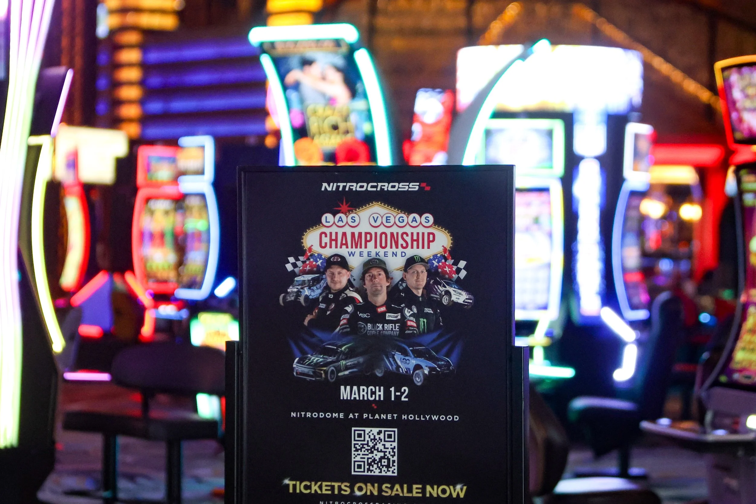 A promotional sign for the NitroCross Las Vegas Championship Weekend event featuring race car drivers, cars, and checkered flags, held at the NitroDome at Planet Hollywood on March 1-2, with bright, colorful arcade machines in the background.