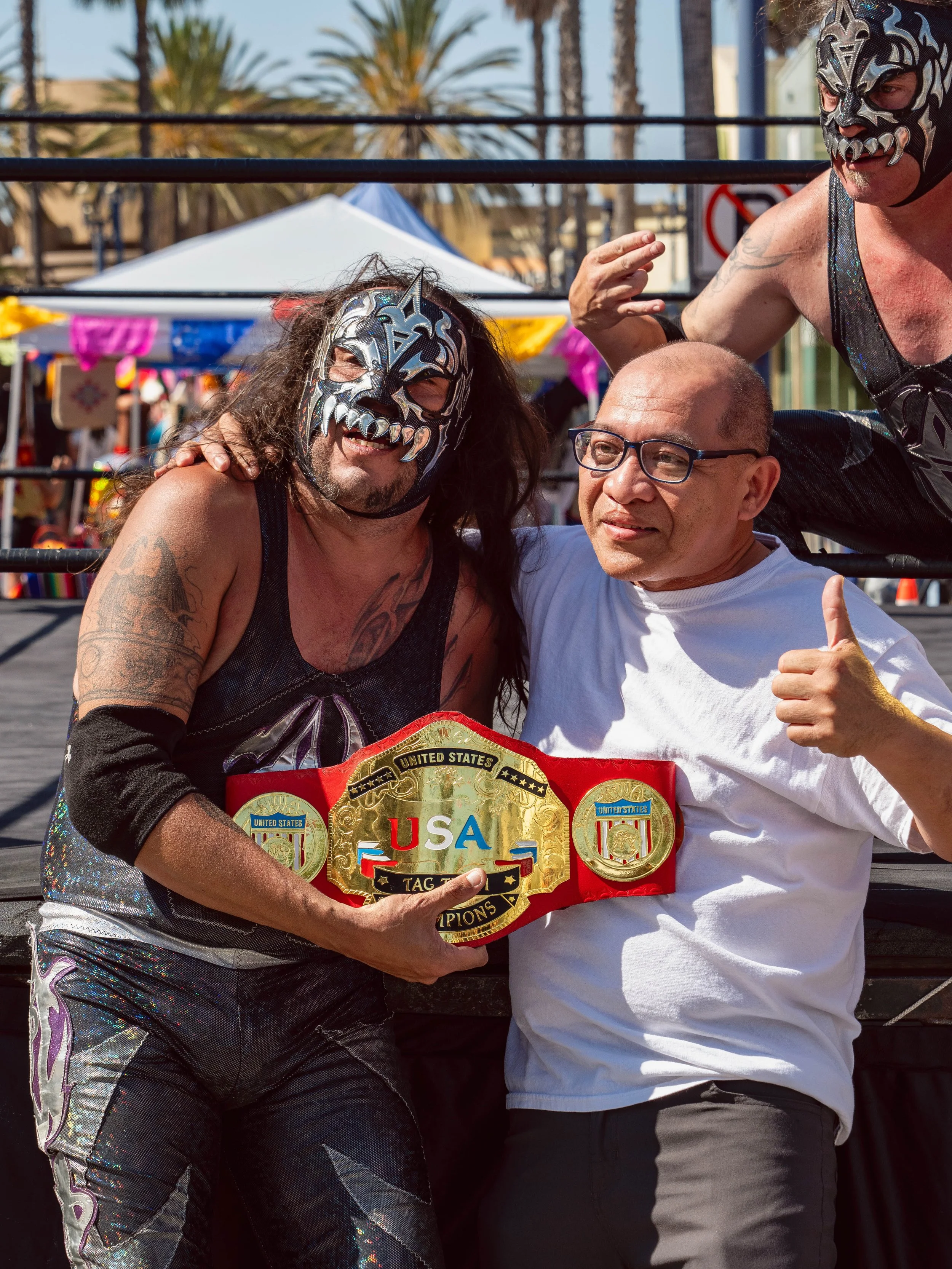 Four people, two wearing lucha libre masks, celebrating with a wrestling championship belt, outdoors in bright sunlight with palm trees and vendor tents in background.