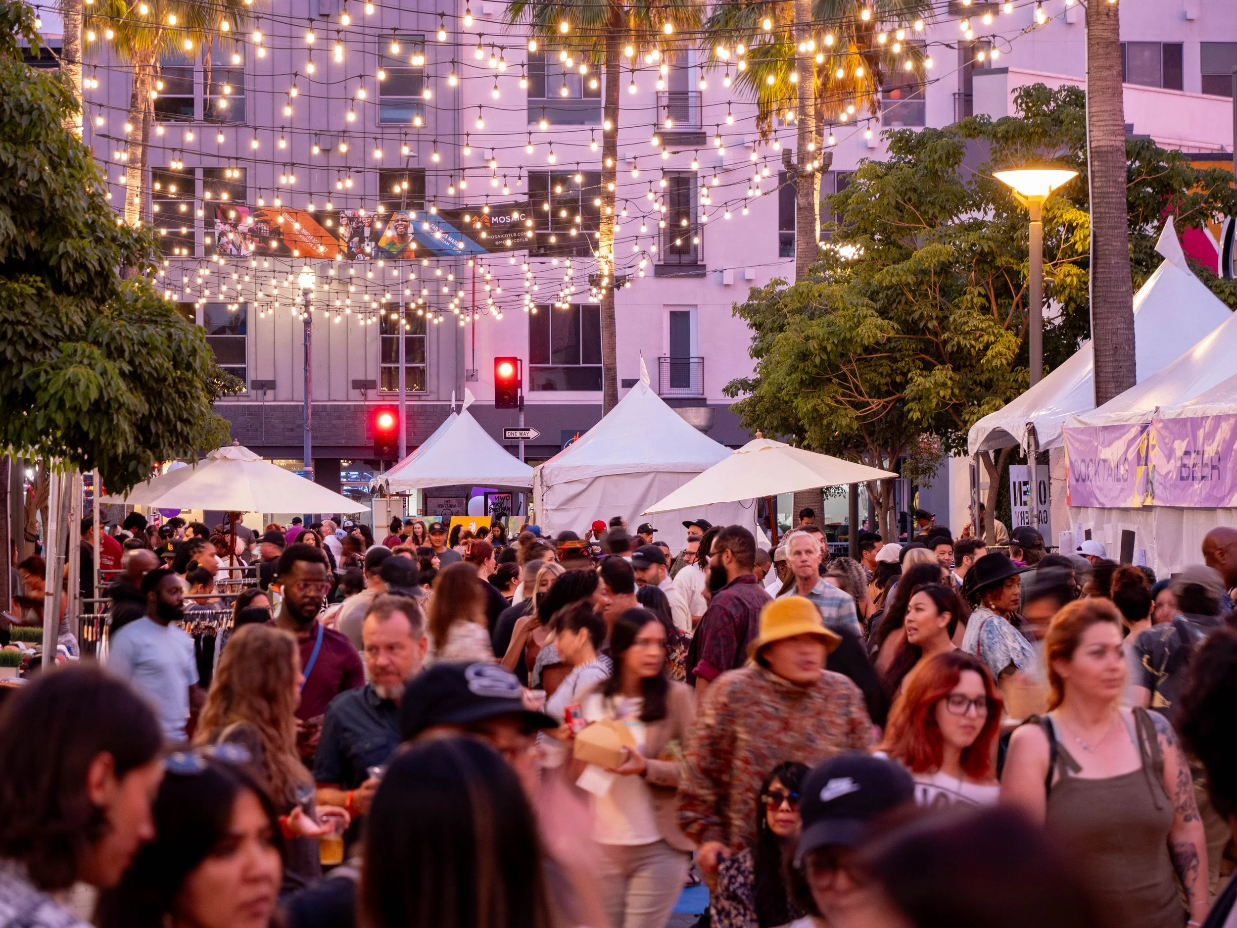 Crowded outdoor street festival at dusk, with people shopping and socializing under string lights and tents, surrounded by trees and city buildings.