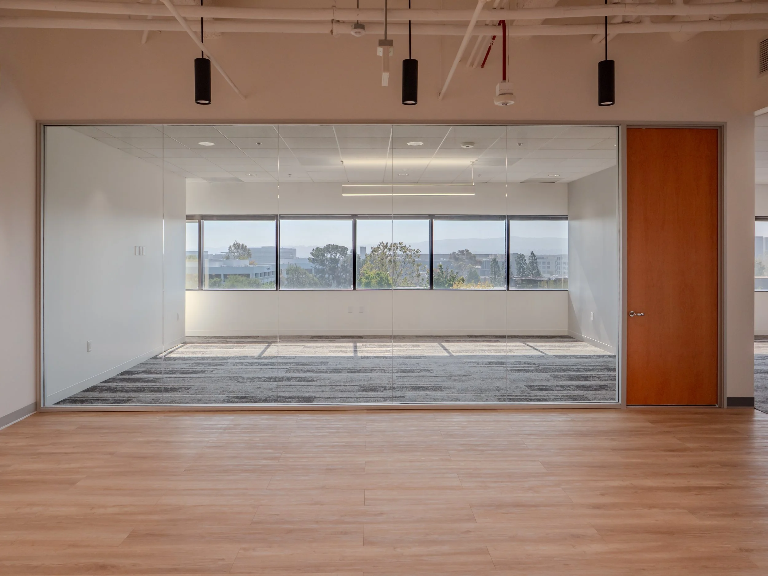 Empty modern office room with wooden flooring, large window wall with a city view, and a glass wall with a wooden door.