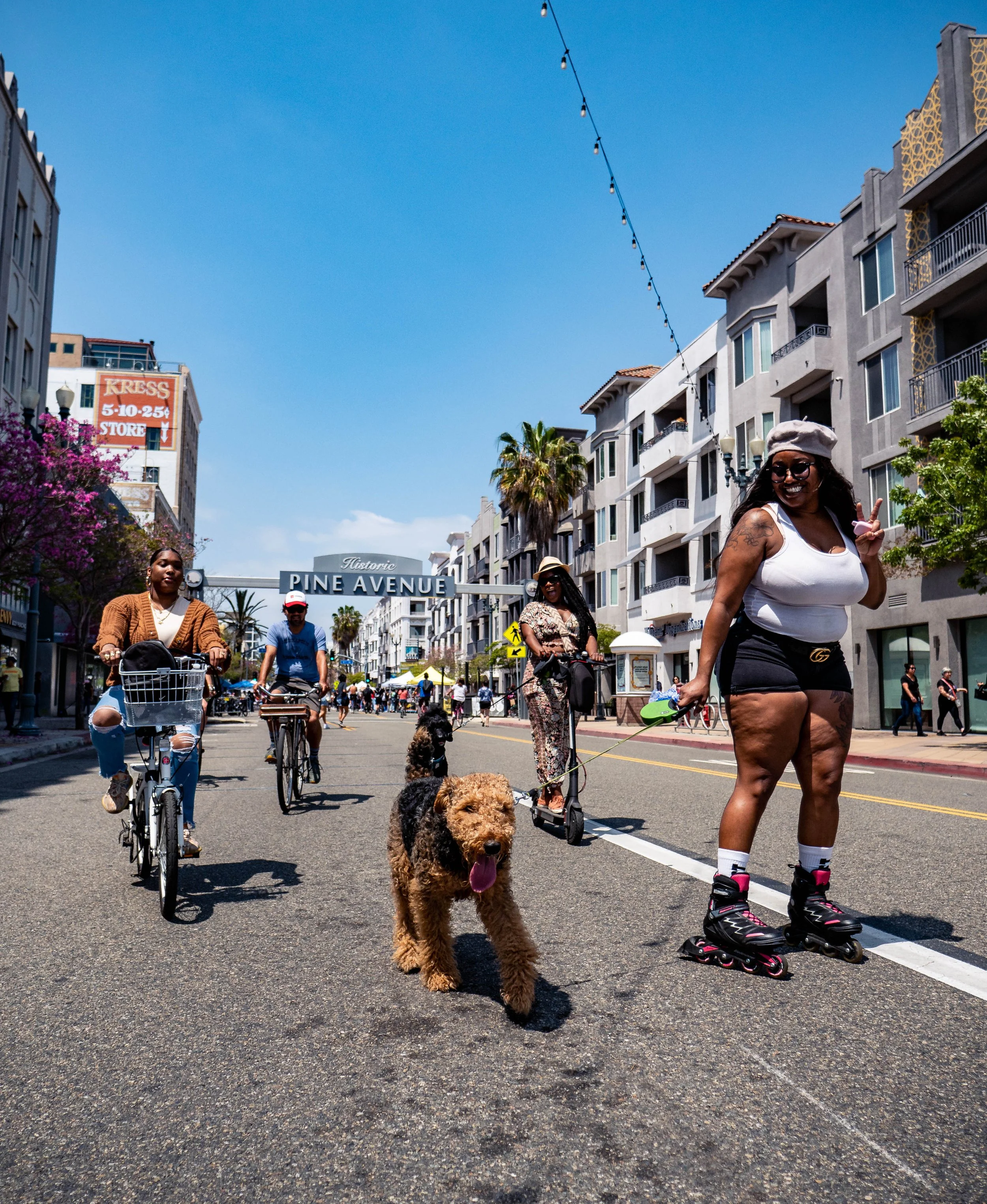People walking and riding bikes in a lively city street, with a woman rollerblading and holding a dog on a leash, under a sign that says 'Historic Pine Avenue.'