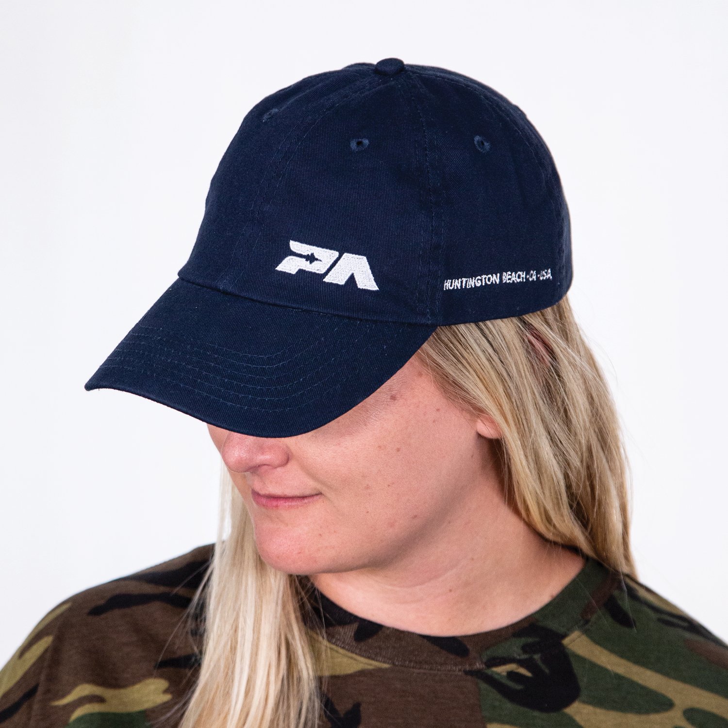 A woman wearing a navy blue cap with white logo and text, dressed in a camouflage shirt, facing slightly downward.