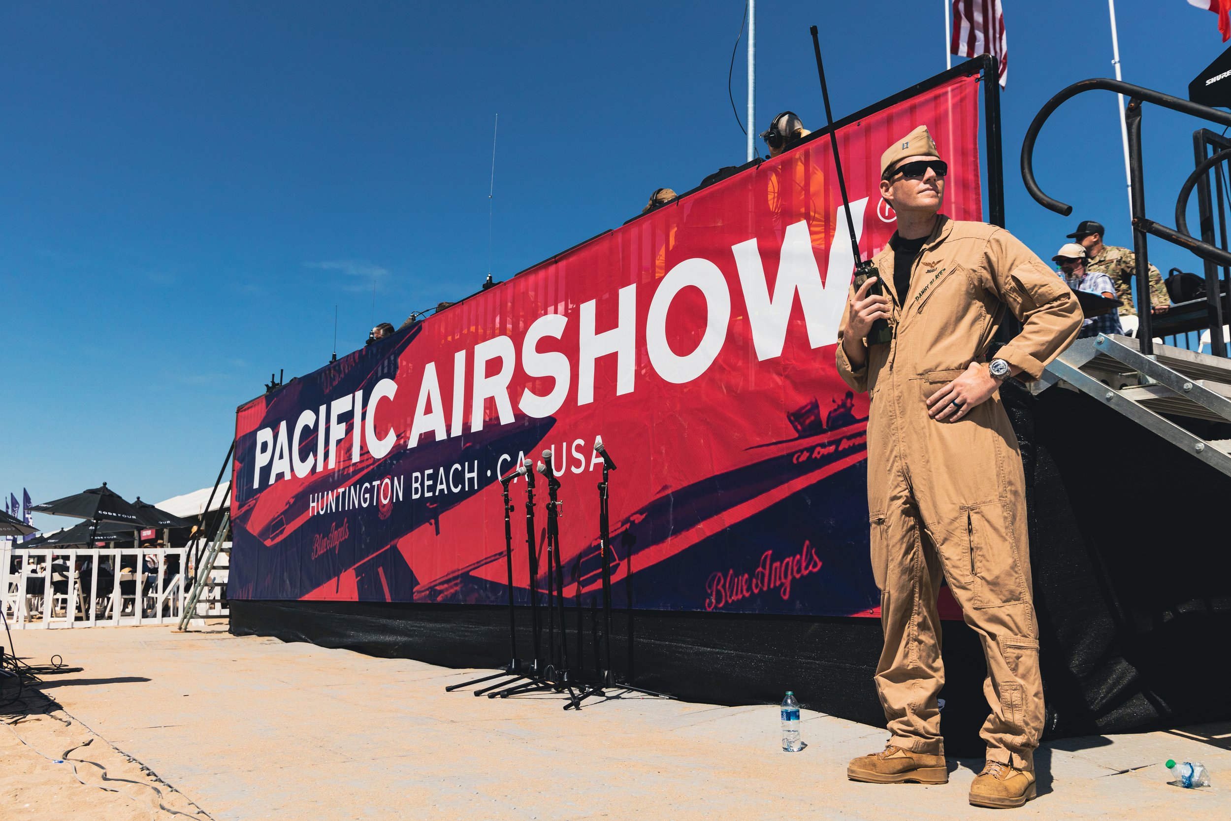 Man in military uniform standing on beach near a large red banner that reads 'Pacific Airshow, Huntington Beach, CA, USA', with several people in the background and clear blue sky.