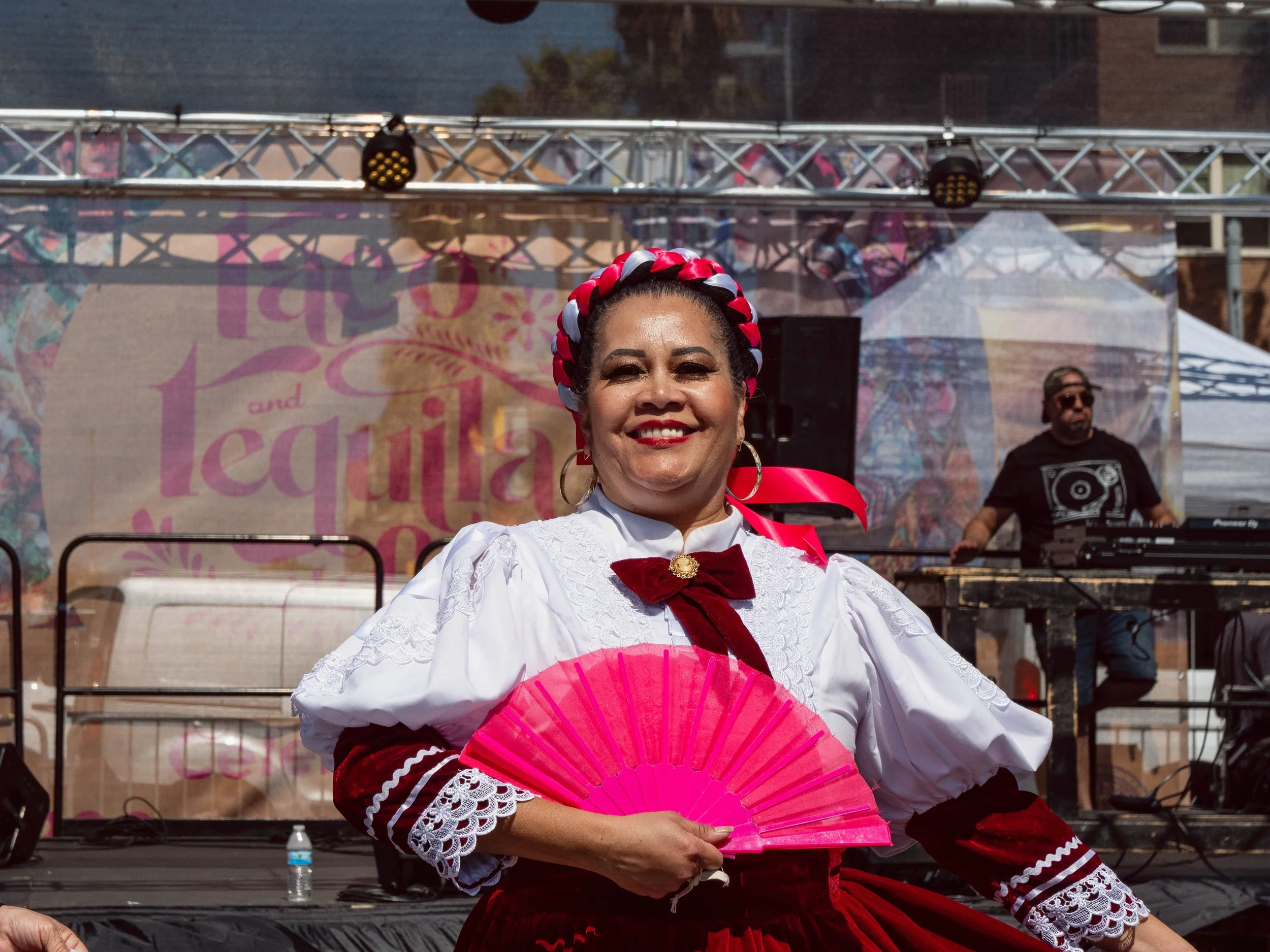 A woman dressed in traditional Mexican attire, holding a pink fan, smiling at an outdoor cultural event with a stage and DJ in the background.