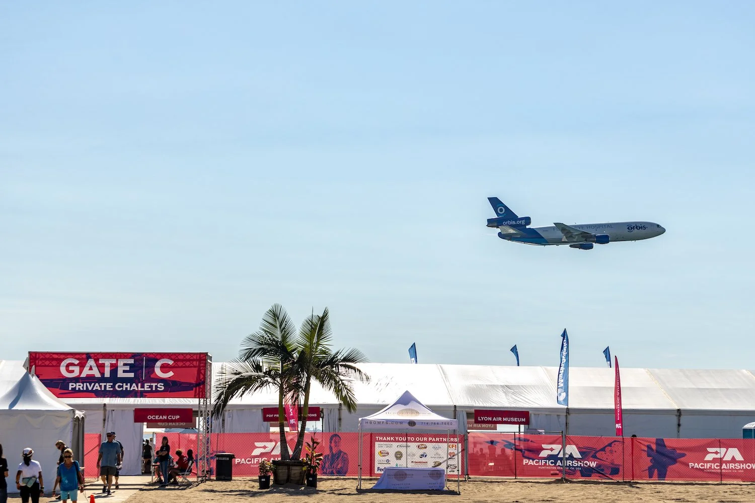 An airplane flying in the sky above a beach festival entrance with palm trees and tents, with signs indicating Gate C and private chalets.