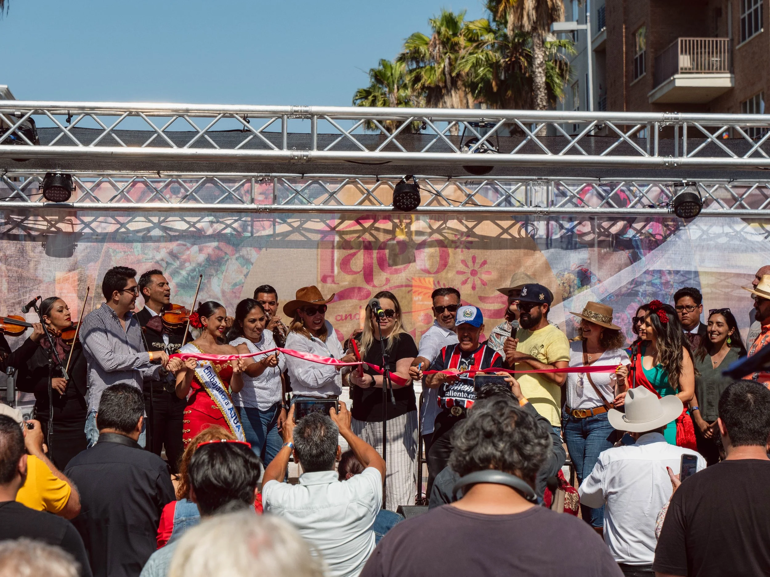 People at a cultural event on stage, some holding a ribbon for a ribbon-cutting ceremony, with musicians playing violins in the background, under sunlight, with palm trees and buildings behind.