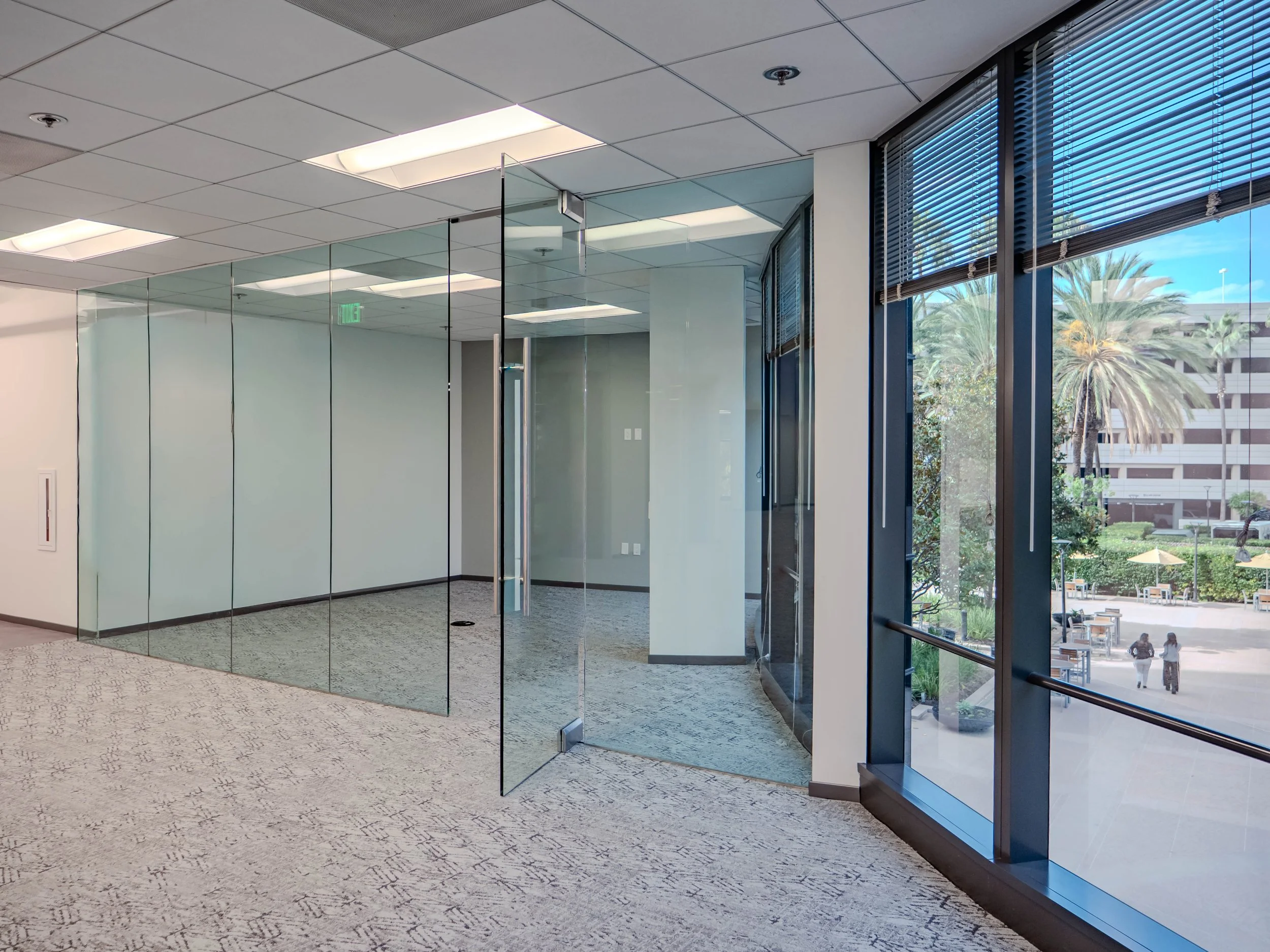 Empty office corner with glass walls and large windows showing outdoor palm trees and a courtyard.