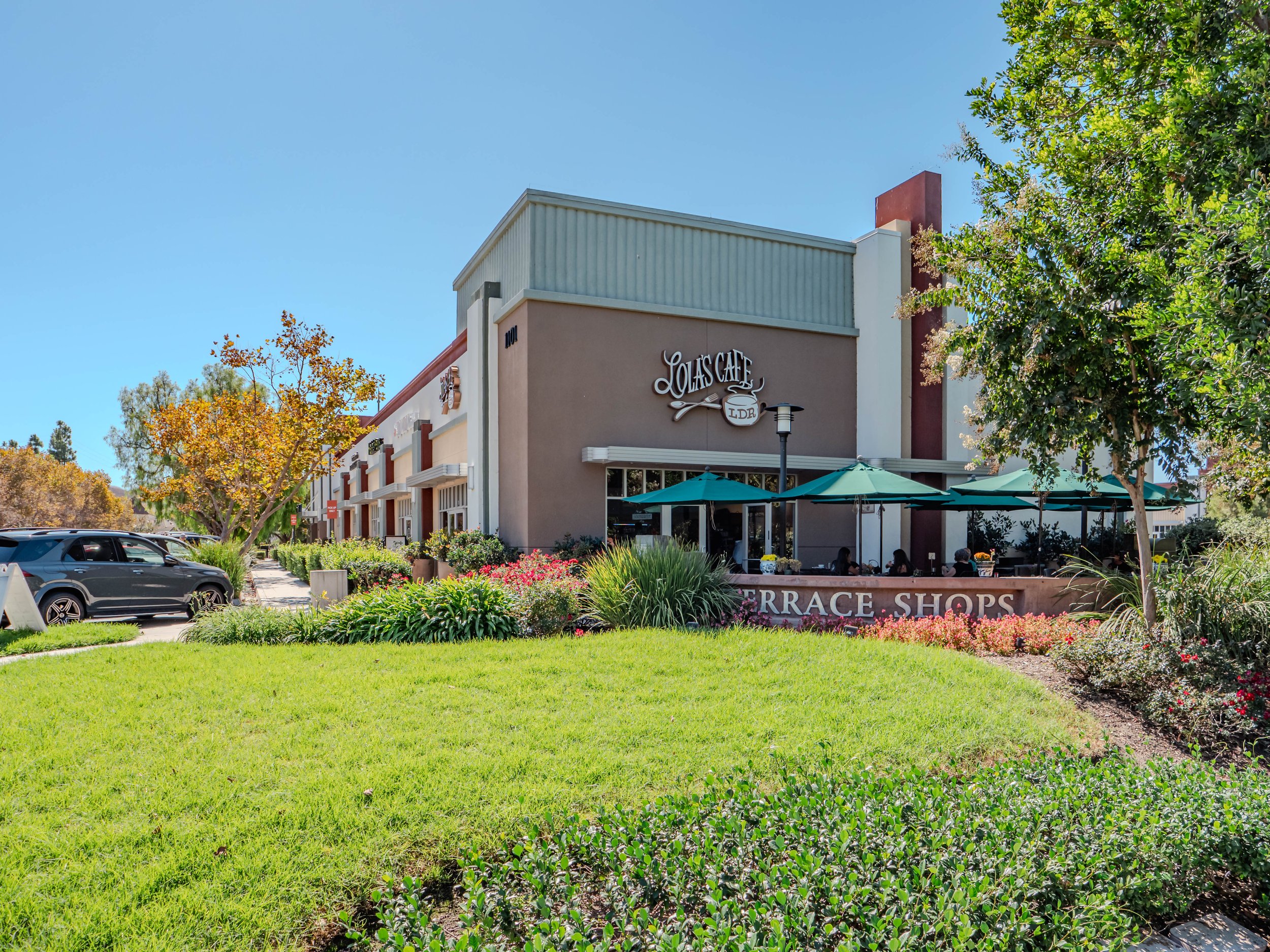 Exterior view of Loa's Cafe with outdoor seating under umbrellas, surrounded by green grass, trees, and colorful bushes, in a shopping plaza with parked cars nearby.