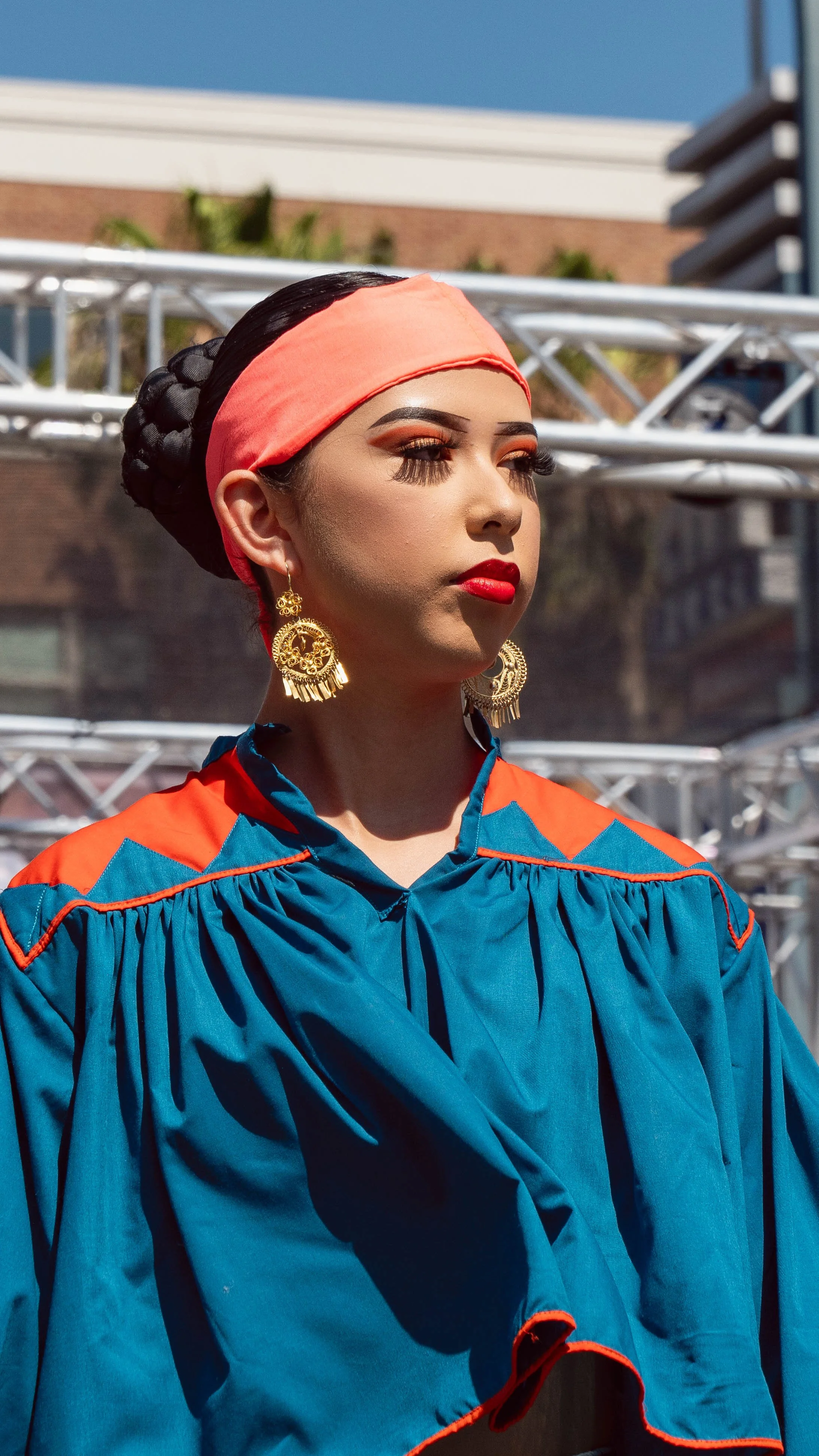 Young woman wearing a blue graduation gown with orange accents, gold earrings, red lipstick, and a coral-colored headscarf, standing outdoors with metal scaffolding and a building in the background.