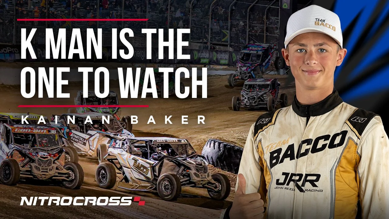 A young male off-road racing driver in a racing suit and cap standing in front of a dirt race track with several off-road vehicles racing on it, promoting K MAN as the one to watch, with the name KAINA BAKER and NitroCross branding.