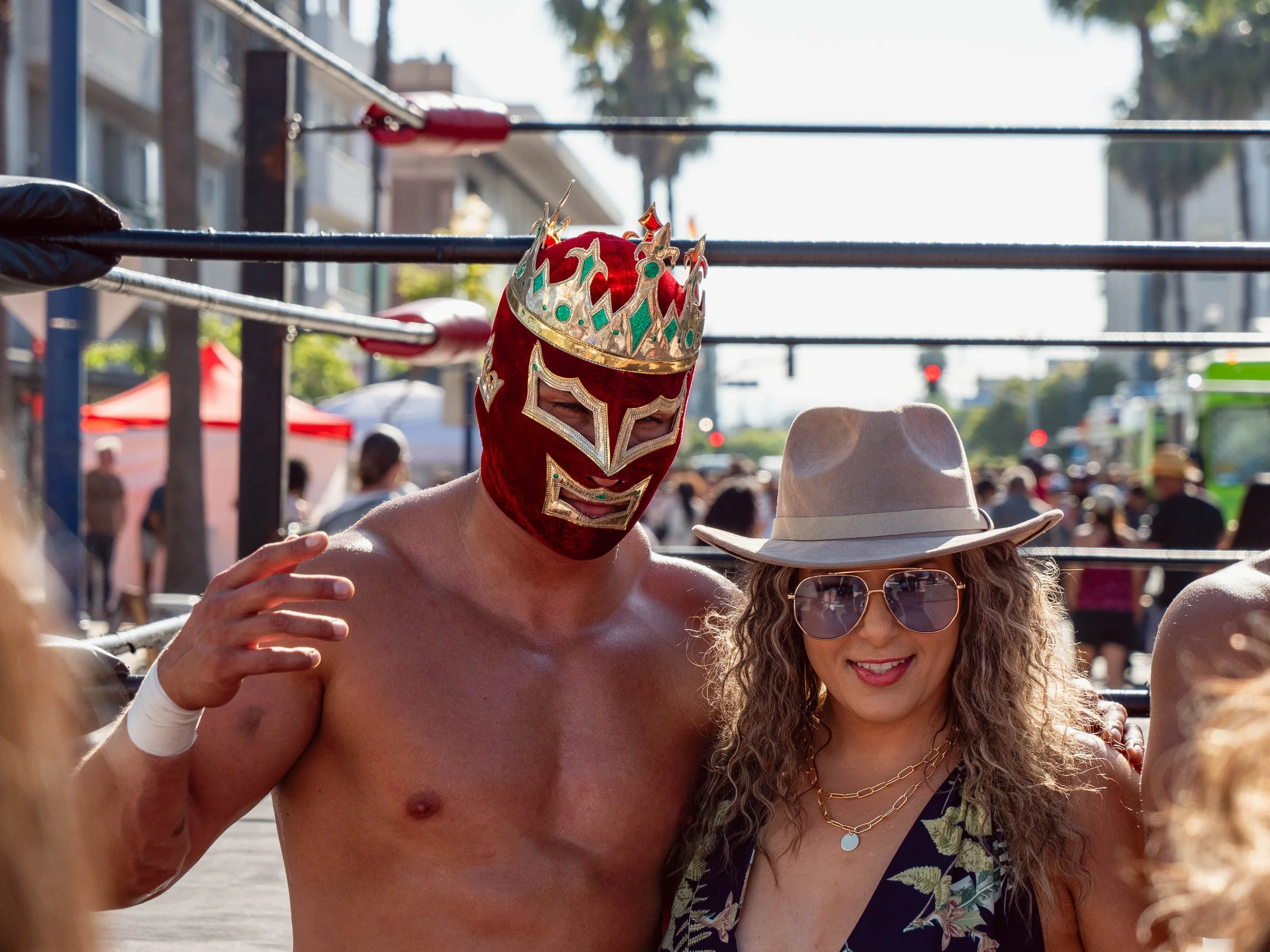 A shirtless man wearing a red luchador mask with gold and green accents, standing next to a woman with long curly hair, sunglasses, and a beige hat, at an outdoor event with a wrestling ring and crowd in the background.