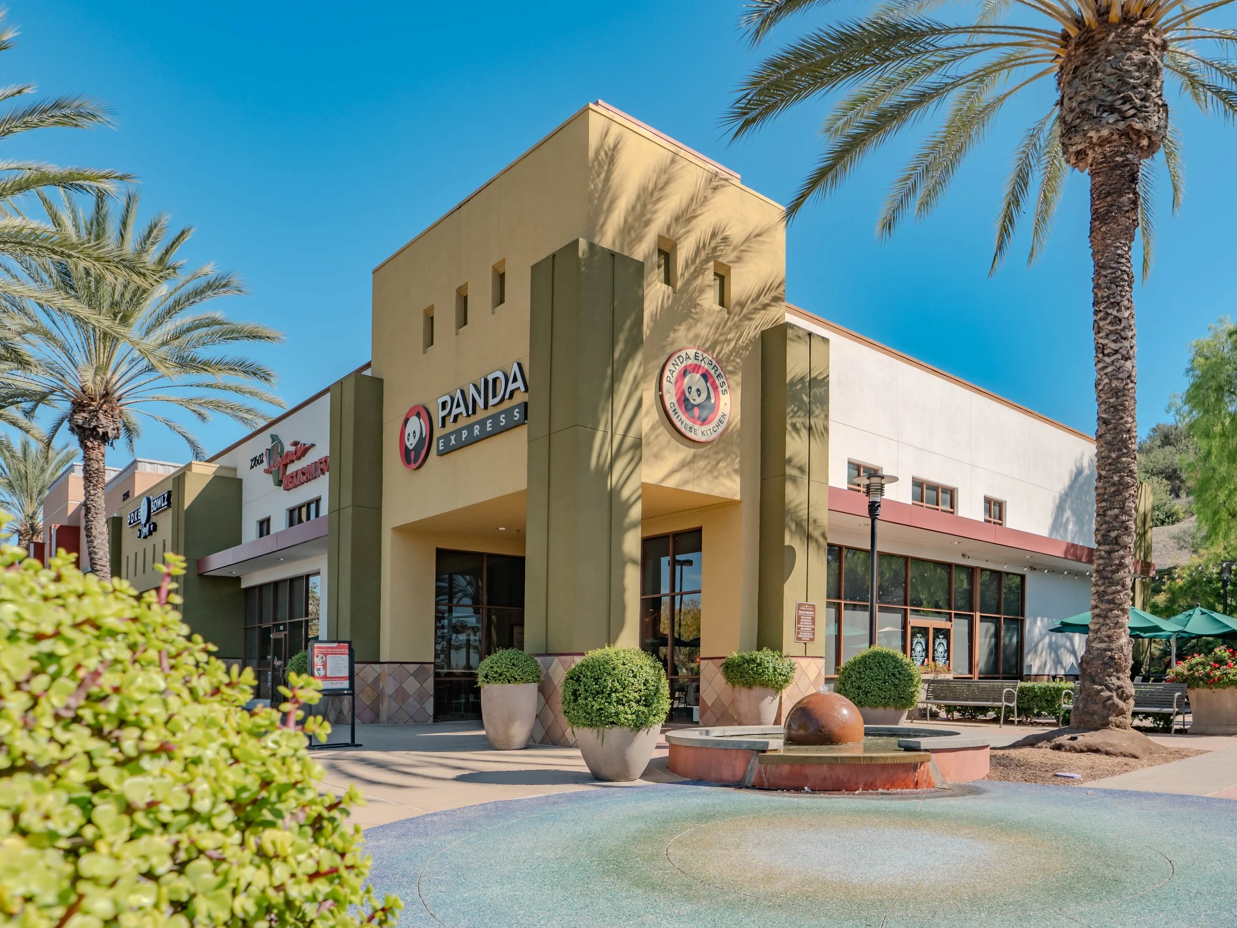 Exterior view of a Panda Express restaurant with palm trees, potted plants, benches, and umbrellas on a sunny day.