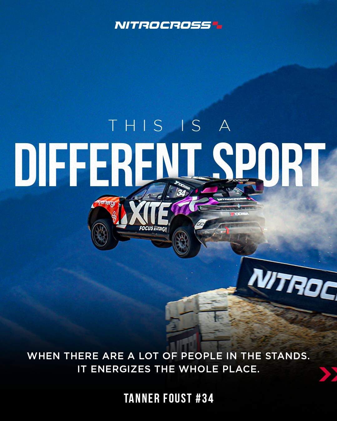 A rally car jumping over a dirt ramp during a NitroCross event, with mountains and a clear blue sky in the background, and NitroCross banners on the side.