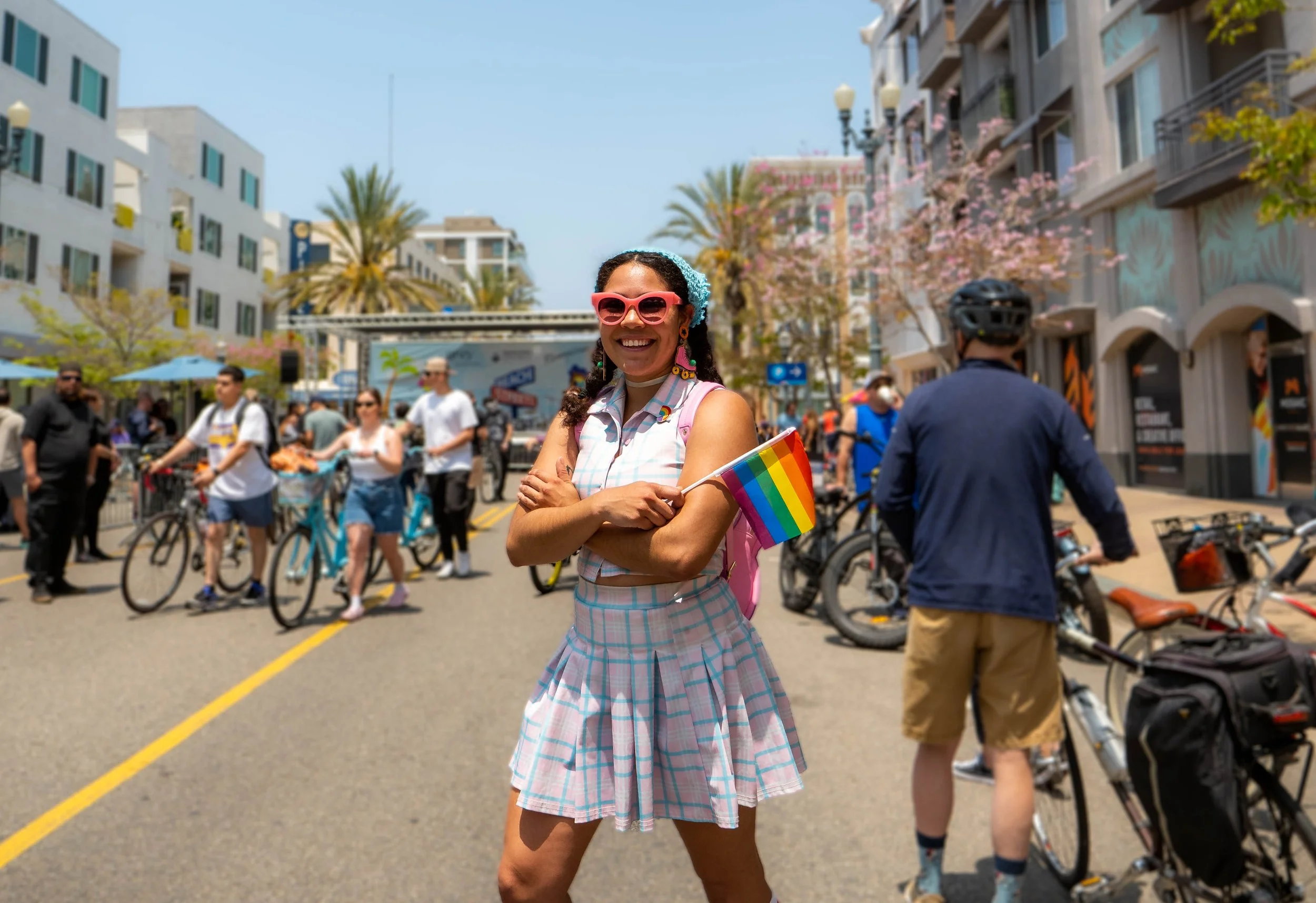A woman smiling at a pride parade holding a rainbow flag, wearing pink sunglasses, a checkered dress, and a blue hair accessory, with a crowd and bicycles in the background.