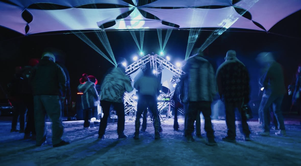 People dancing in front of a DJ booth with colorful laser lights and a canopy overhead at a nightclub or music event.