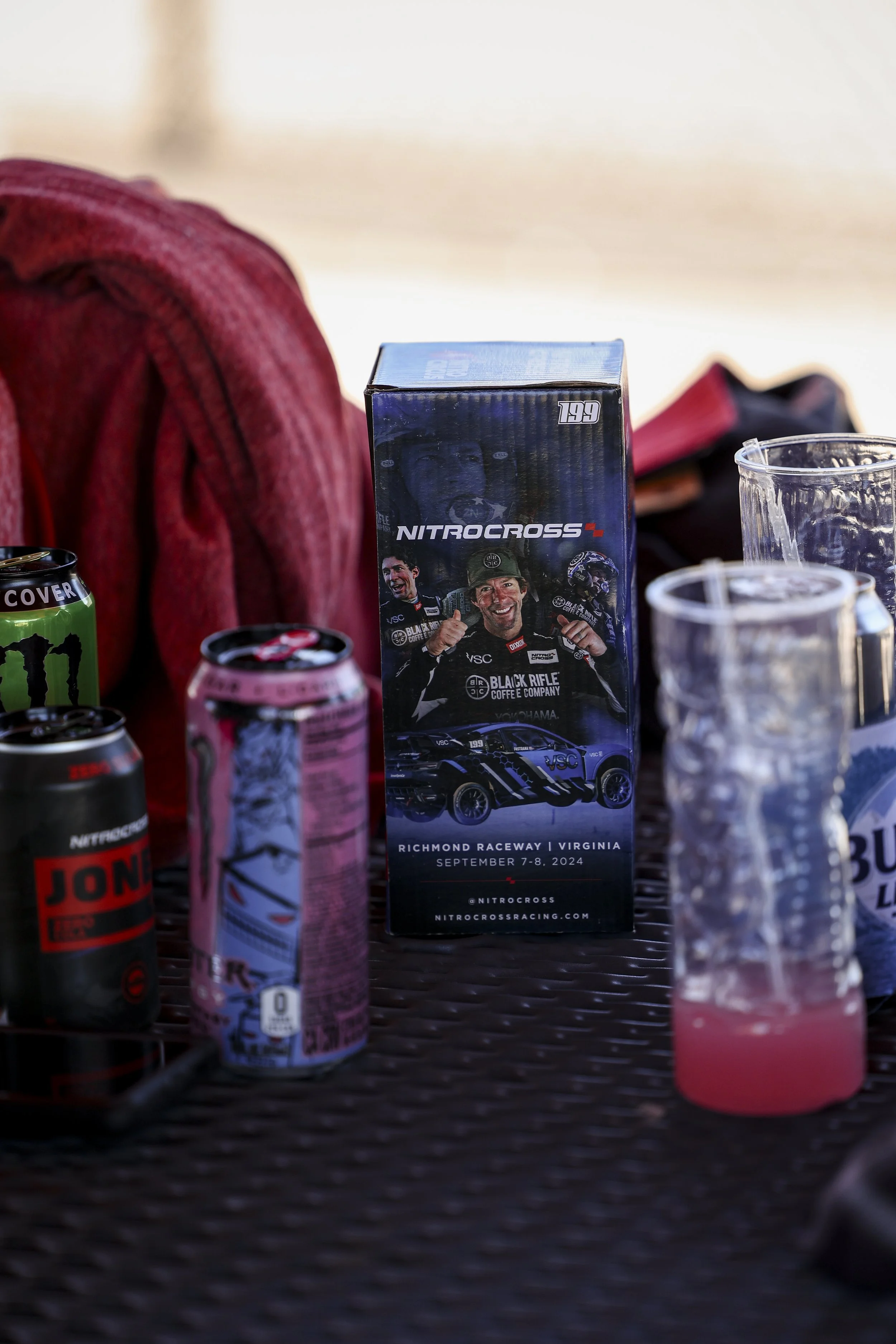 A table with various cans and cups, featuring a NitroCross racing event flyer for September 7-8, 2024 at Richmond Raceway, Virginia, with images of race car drivers and a race car.
