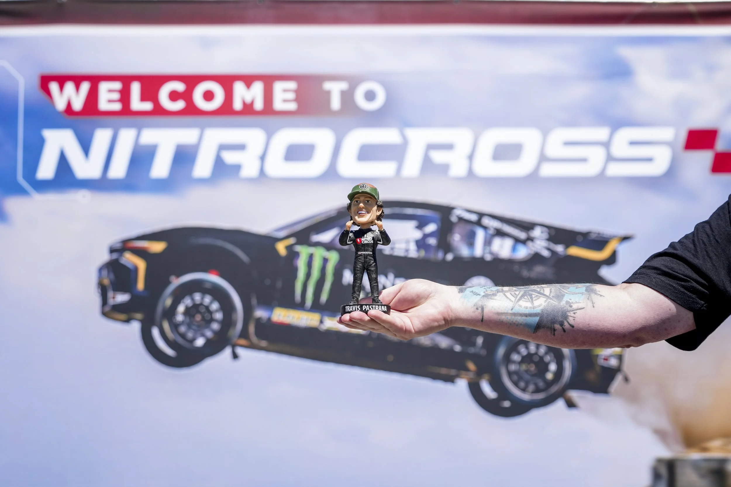 A hand holding a Travis Pastrana bobblehead figurine in front of a Nitro Cross sign and a black race car with Monster Energy logo.