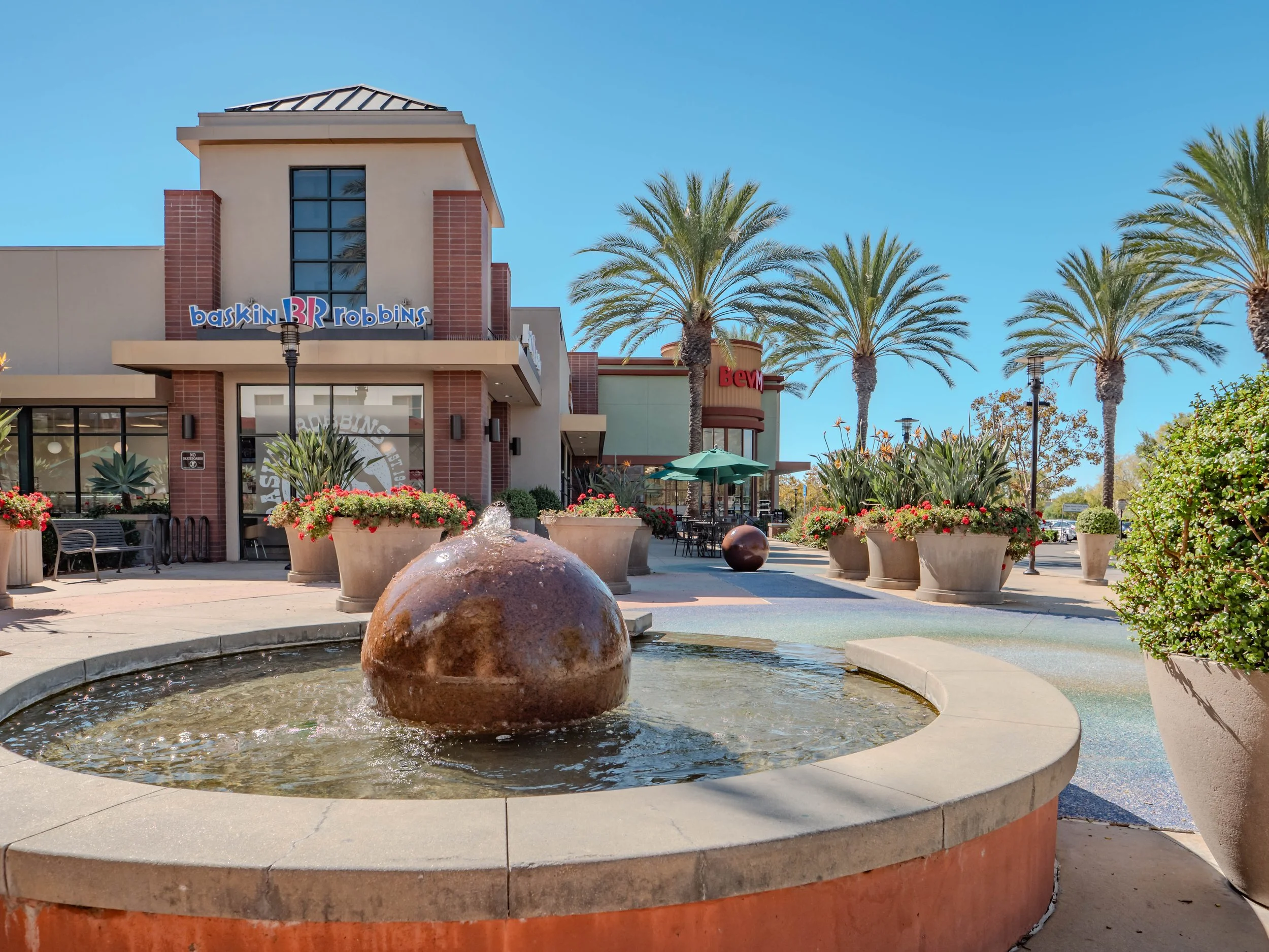 Shopping plaza with a fountain in the foreground, benches, planters with flowers, palm trees, and storefronts including Baskin Robbins and BEVM on a sunny day.