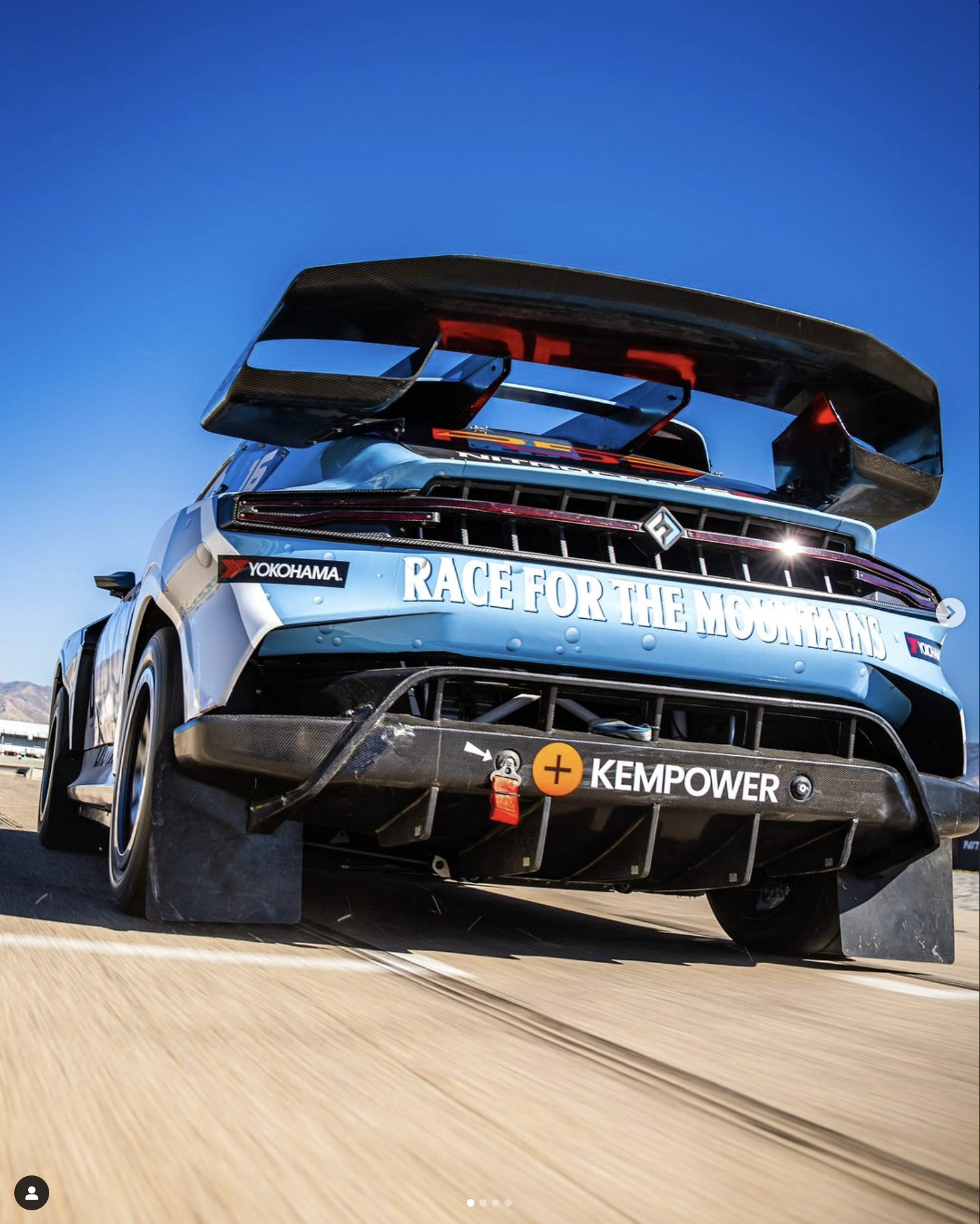 Close-up of race car from behind on a racetrack with a mountain in the background, featuring a large rear wing and sponsorship decals.