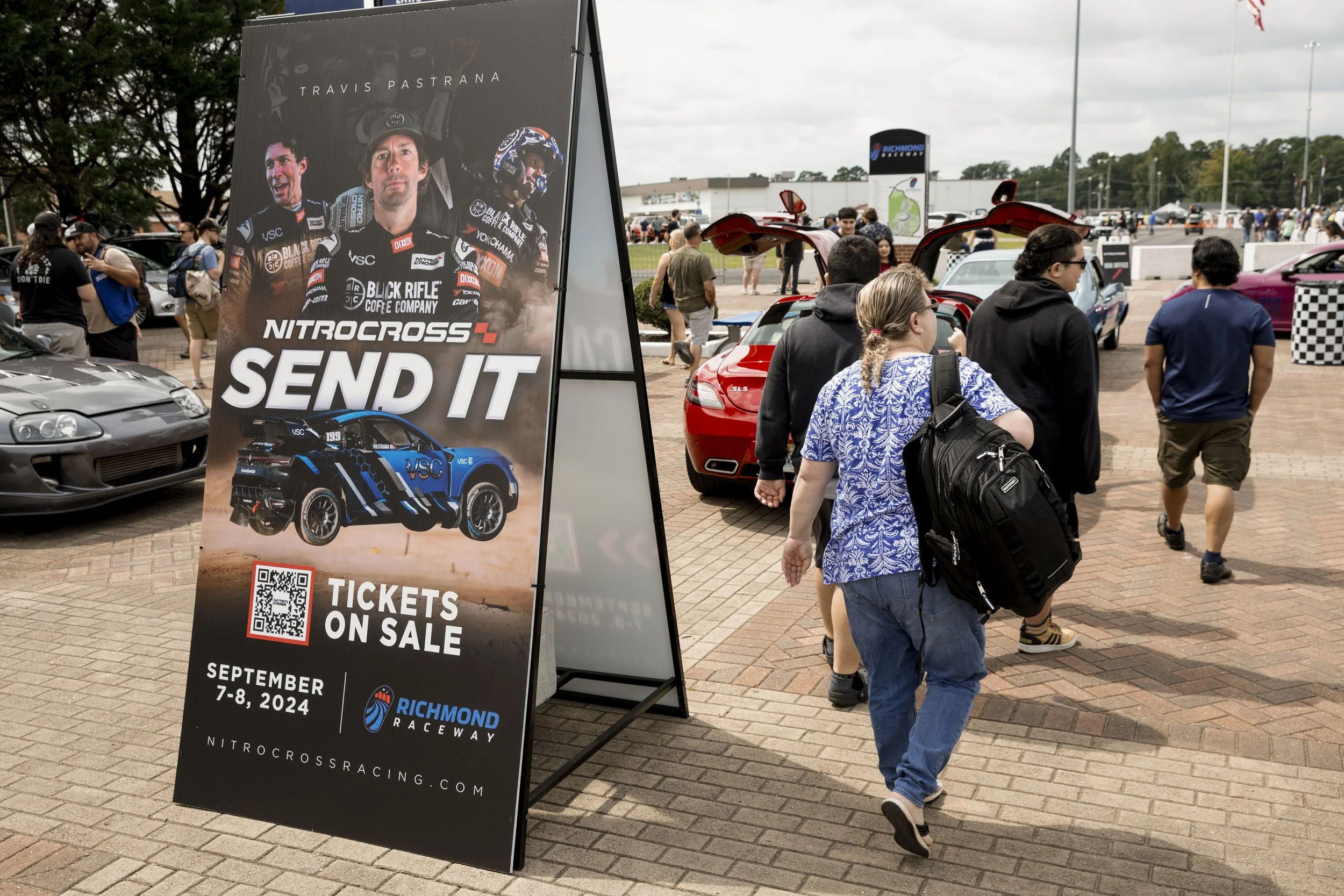 People walking past a large sign advertising NitroCross race tickets for September 7-8, 2024 at Richmond Raceway, with cars on display and an event crowd in the background.