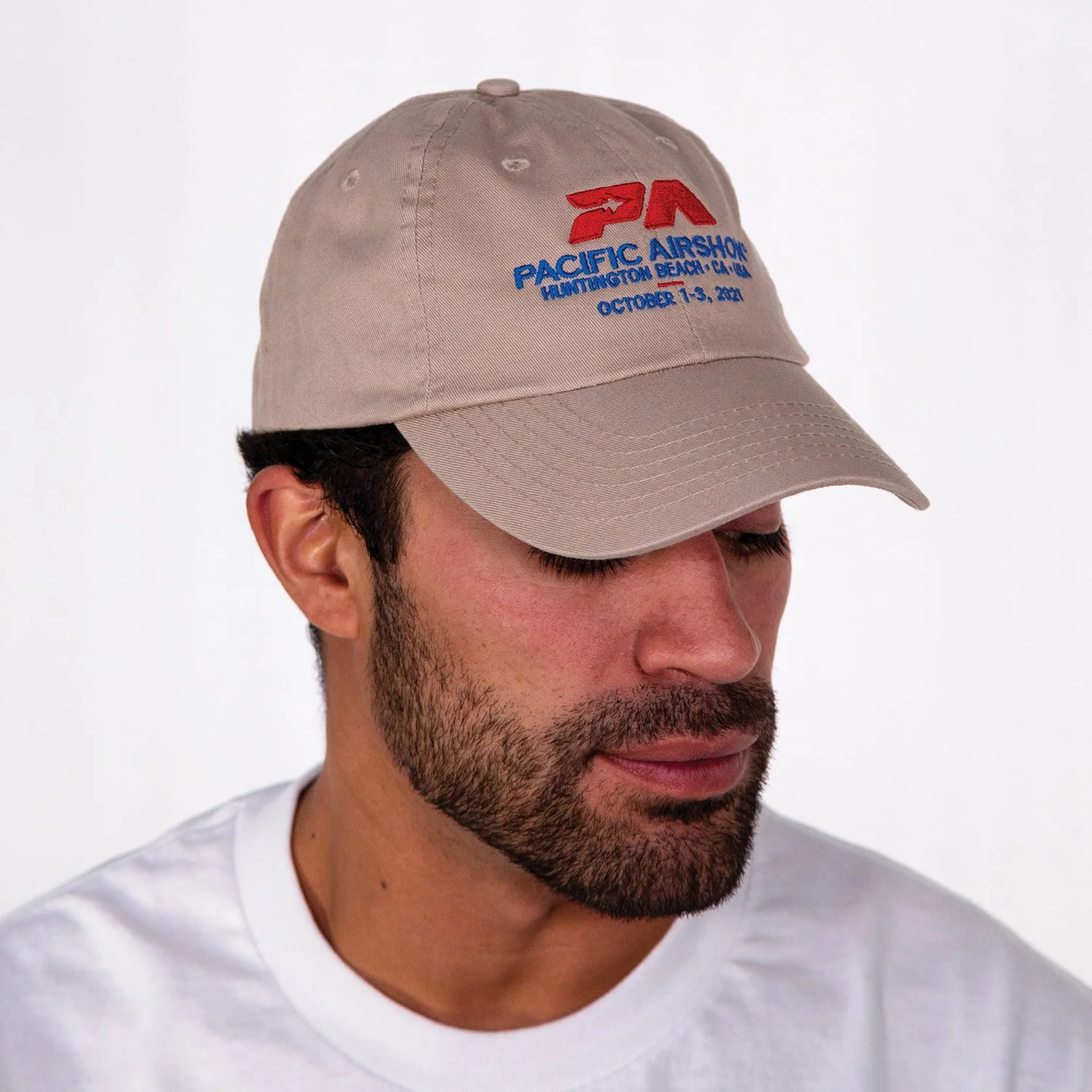 Man with dark hair and beard wearing a beige baseball cap with Pacific Airshow logo and text, and a white T-shirt, looking downward, against a plain white background.