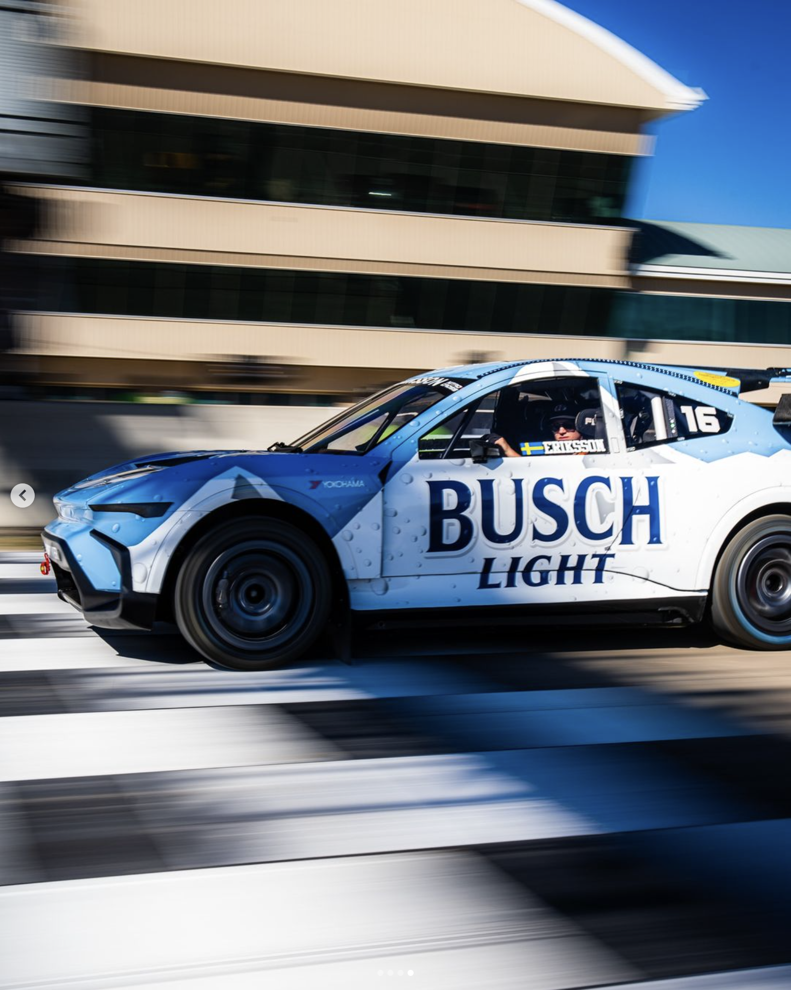 A race car with blue and white racing livery speeding on a track, blurred background, large Busch Light logo on the side, driver wearing a helmet in the cockpit.