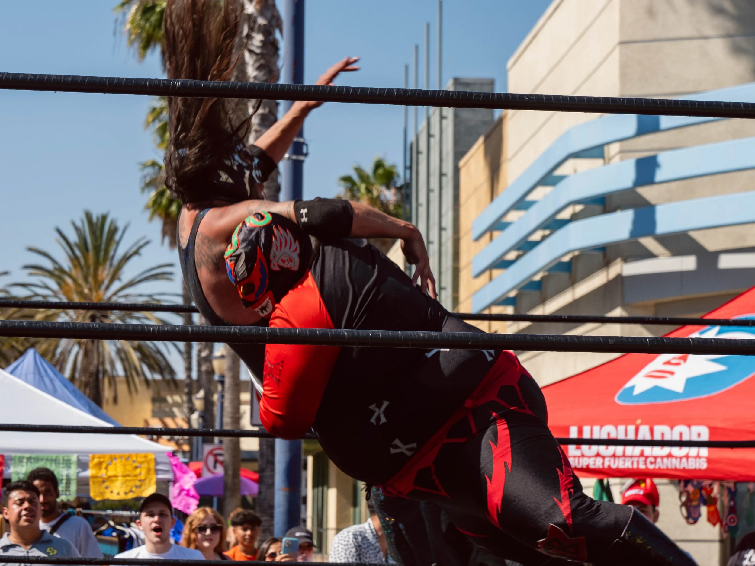 A person with long hair wrestling in a ring during a public event, with spectators and palm trees in the background on a sunny day.