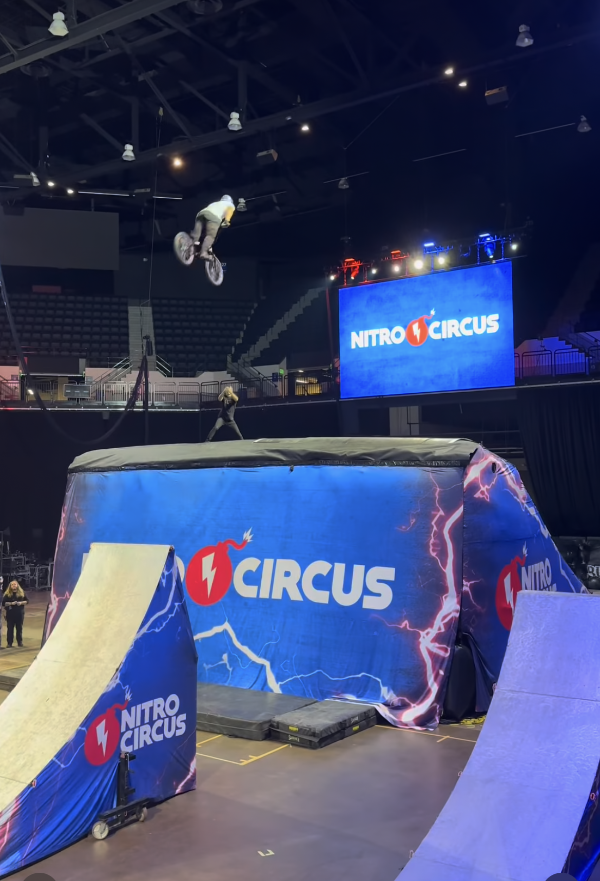 A Nitro Circus stunt show with a motorcyclist performing a jump into an airbag. The arena has a large screen displaying the Nitro Circus logo, and the area is equipped with ramps and safety padding.