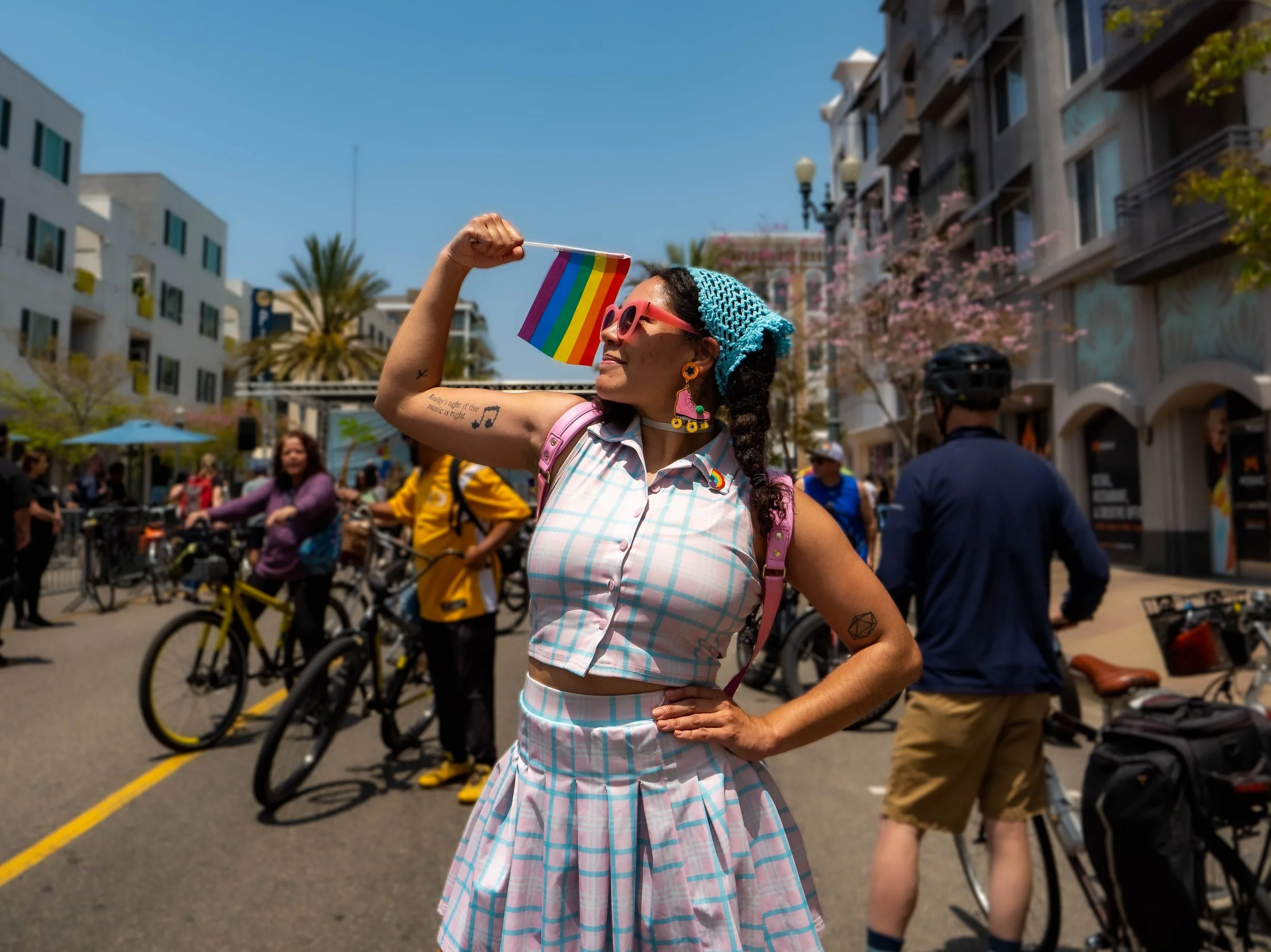 A woman at an outdoor event holds a rainbow pride flag and wears pink sunglasses, a blue headscarf, and a pastel checkered outfit, surrounded by people with bicycles on a city street.