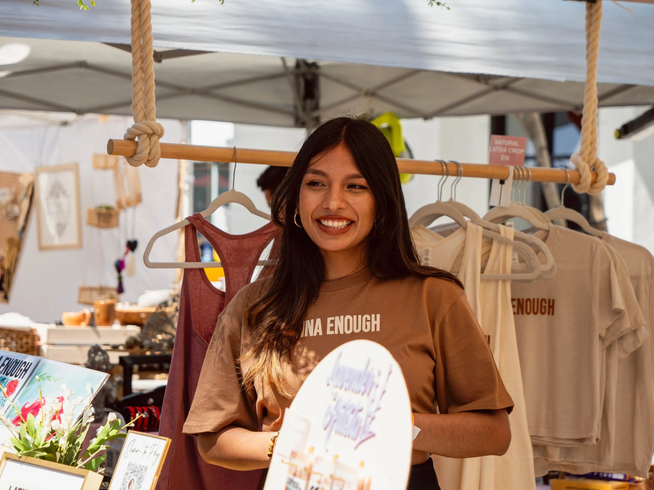 A smiling woman with long dark hair and hoop earrings standing in front of a clothing stall at an outdoor market. The stall has beige and rust-colored clothing on display, with a sign that reads 'NATURAL COTTON' in the background.