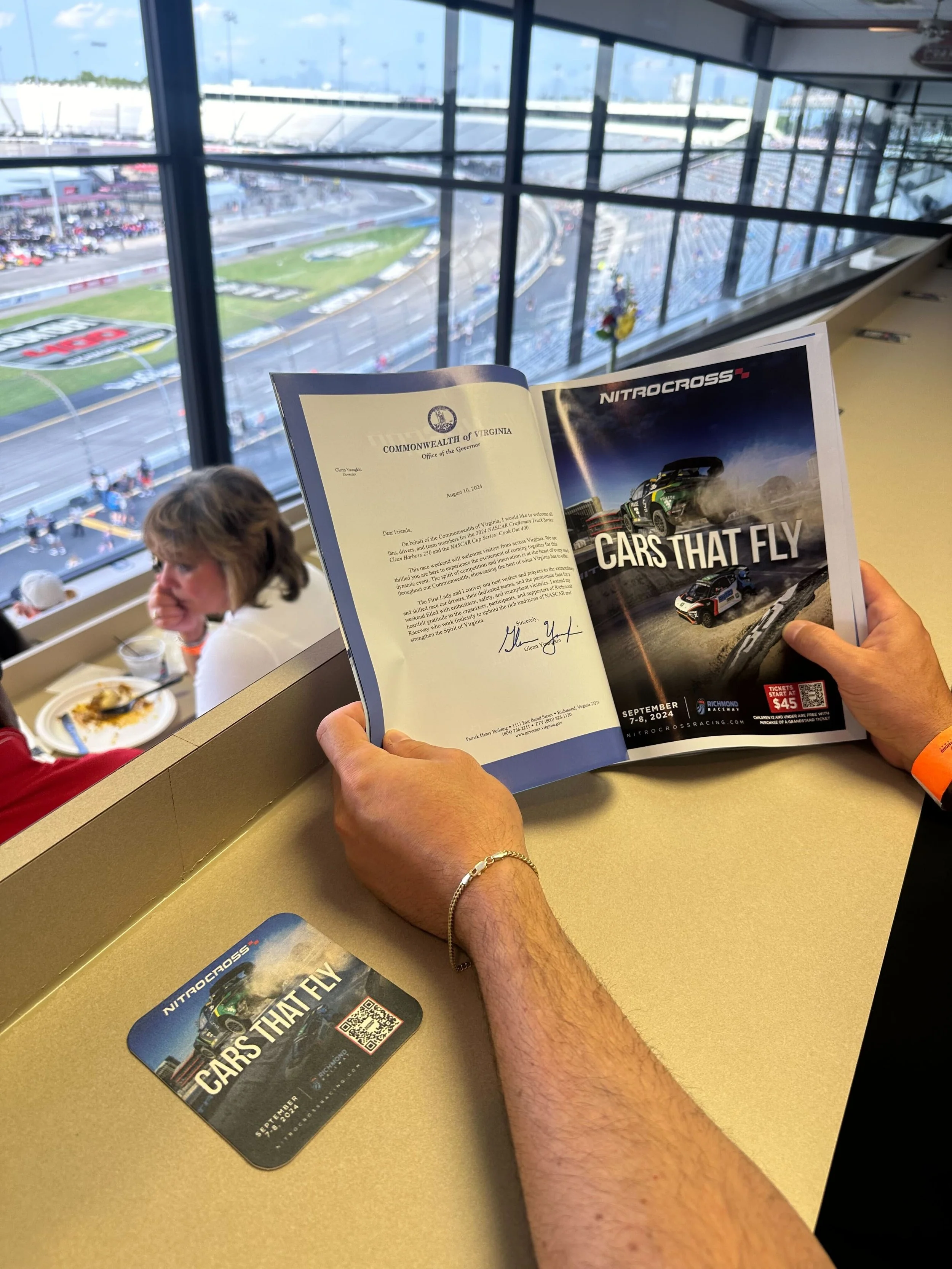 Person holding a NitroCross Racing event brochure with tickets on a restaurant counter at Richmond Raceway, overlooking a race track.