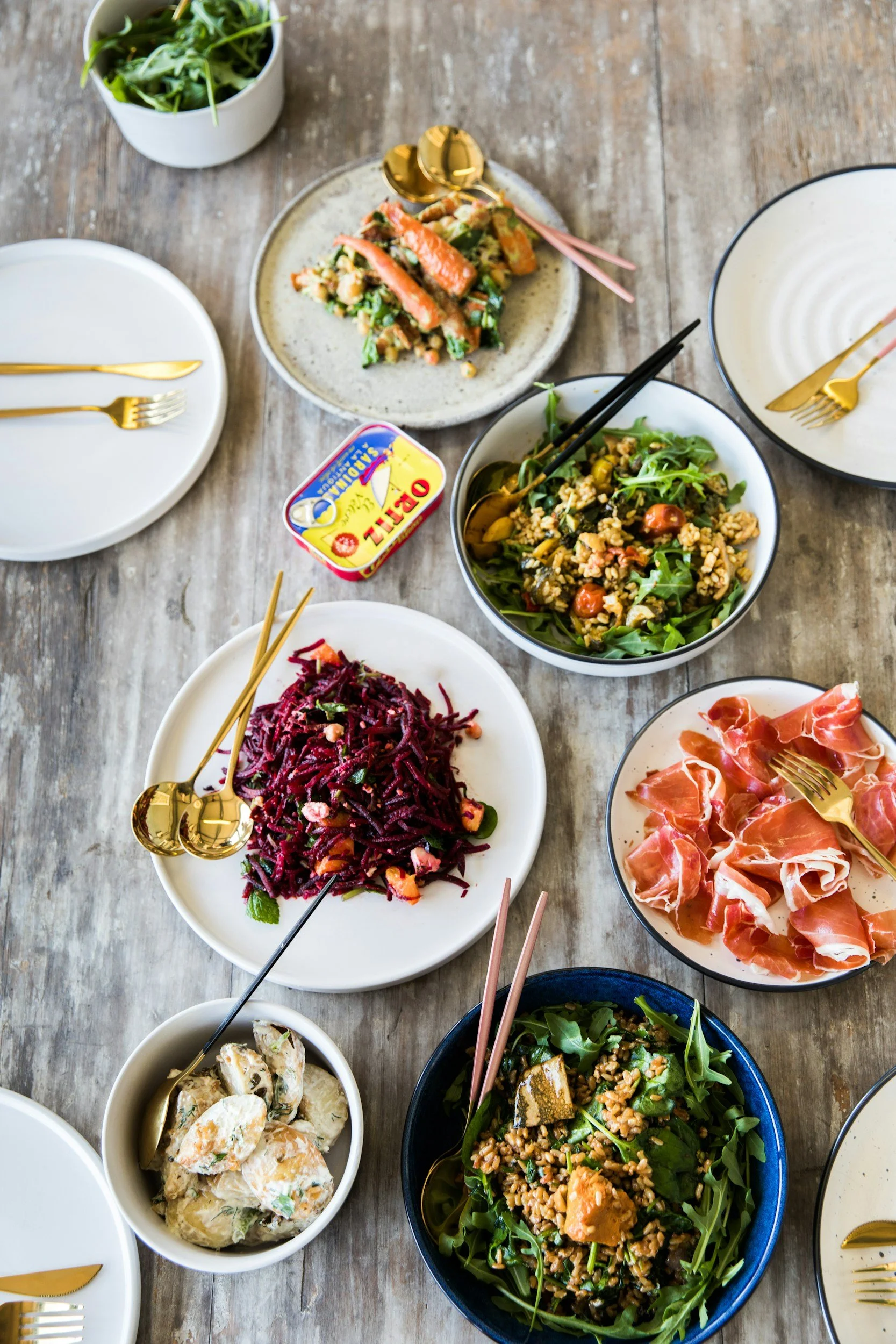 A table set with various salads, a small bowl of potato salad, a container of butter, and bowls of greens. Gold and pink utensils are also on the table.